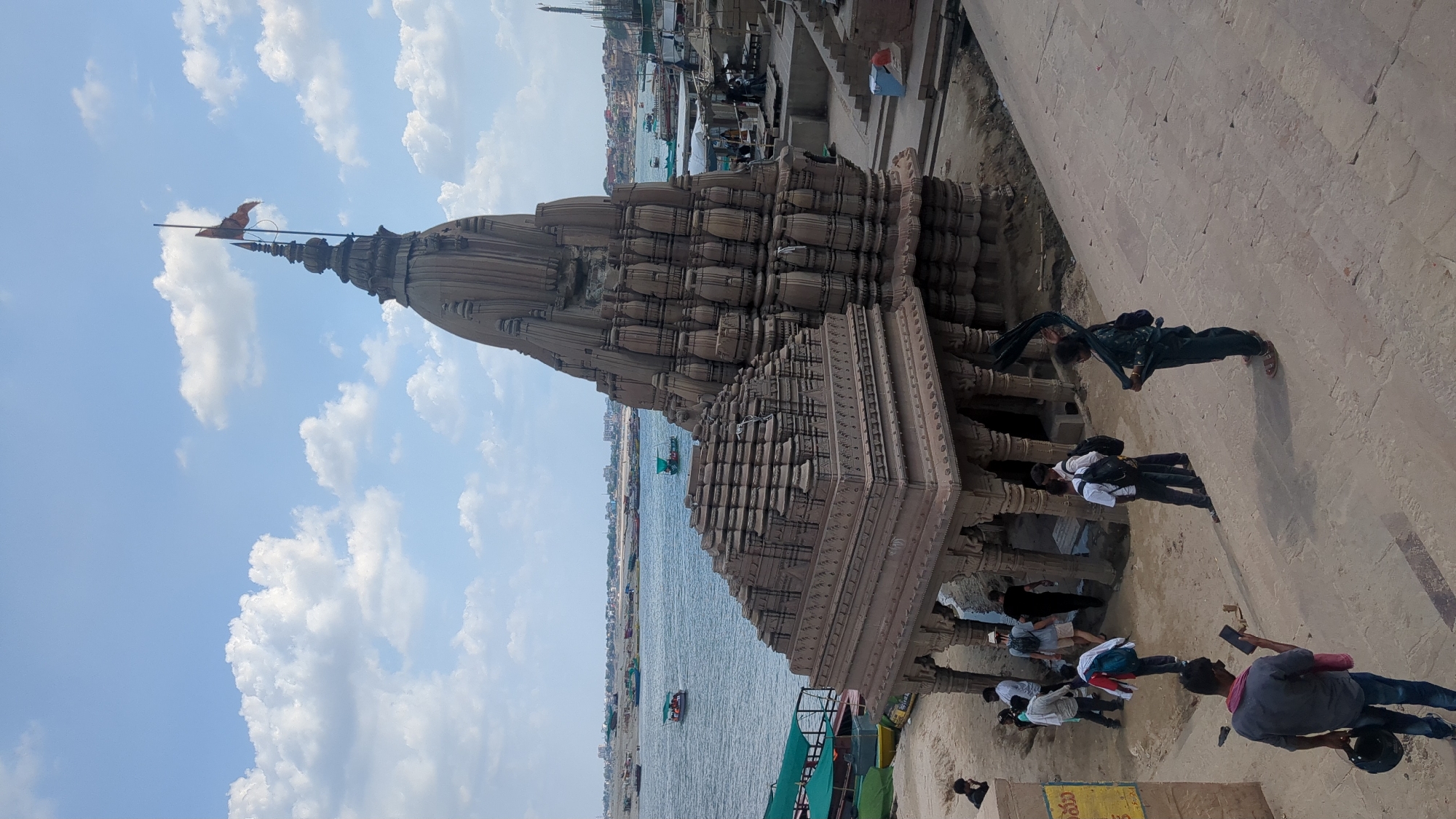 This image depicts the historic Batuk Bhairav Temple complex situated on the banks of the Ganges River in the city of Varanasi, India. The temple features intricate stone carvings and a tall, tiered spire, topped with a flag. In the foreground, several individuals are seen walking on a concrete staircase, some carrying bags. A man in a gray shirt holds a phone and a helmet, while a woman in dark clothing holds a flowing scarf. In the middle ground, a group of people are gathered near the temple entrance, and more figures are visible inside the temple's open structure. The background shows the vast Ganges River with numerous boats, and the distant cityscape of Varanasi. The sky is partly cloudy, with large white clouds against a blue backdrop. The overall scene captures a moment of activity and cultural significance at a prominent religious site.