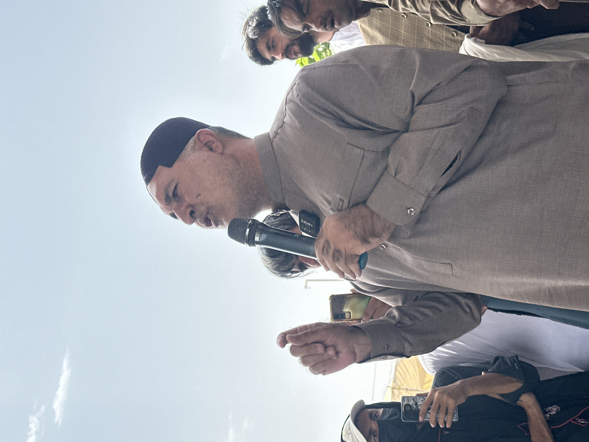 In Quetta City Tehsil, Pakistan, a man wearing a dark skullcap and a light brown tunic speaks into a microphone, gesturing with his left hand. He is surrounded by other men, some of whom are holding cell phones and looking towards him. The background is a pale blue sky with a wispy cloud. The overall scene appears to be an outdoor gathering or event.