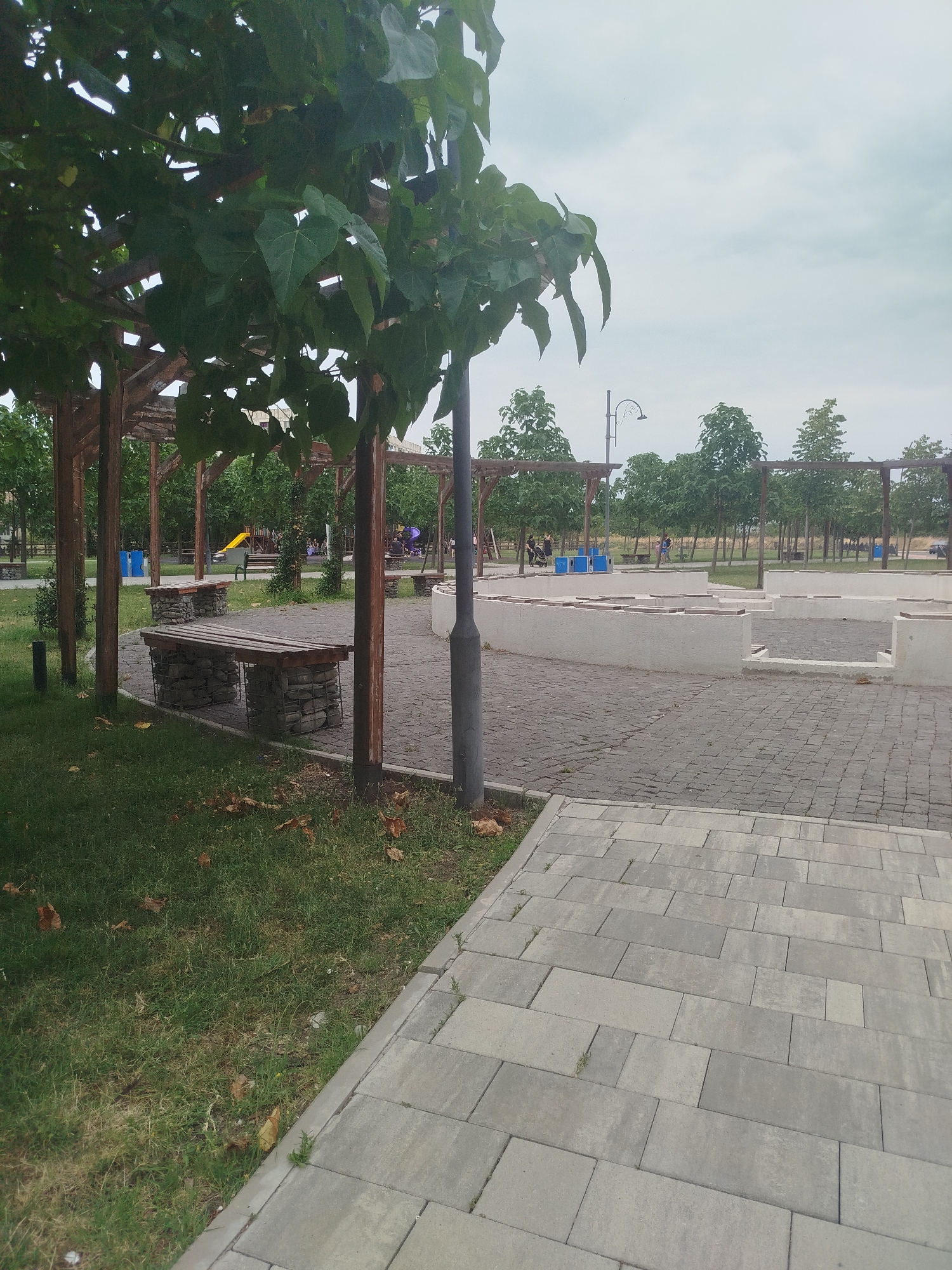 The image shows an outdoor public space with paved walkways and grassy areas. There are several wooden pergolas with green foliage providing shade. Benches made of wood and stone are visible. In the background, there is a playground with a yellow slide and several trees lining the perimeter of the area. Some people are walking in the distance. The sky is overcast. This scene is located in Rustavi, Georgia, specifically in the park area near the Rustavi Steel plant.