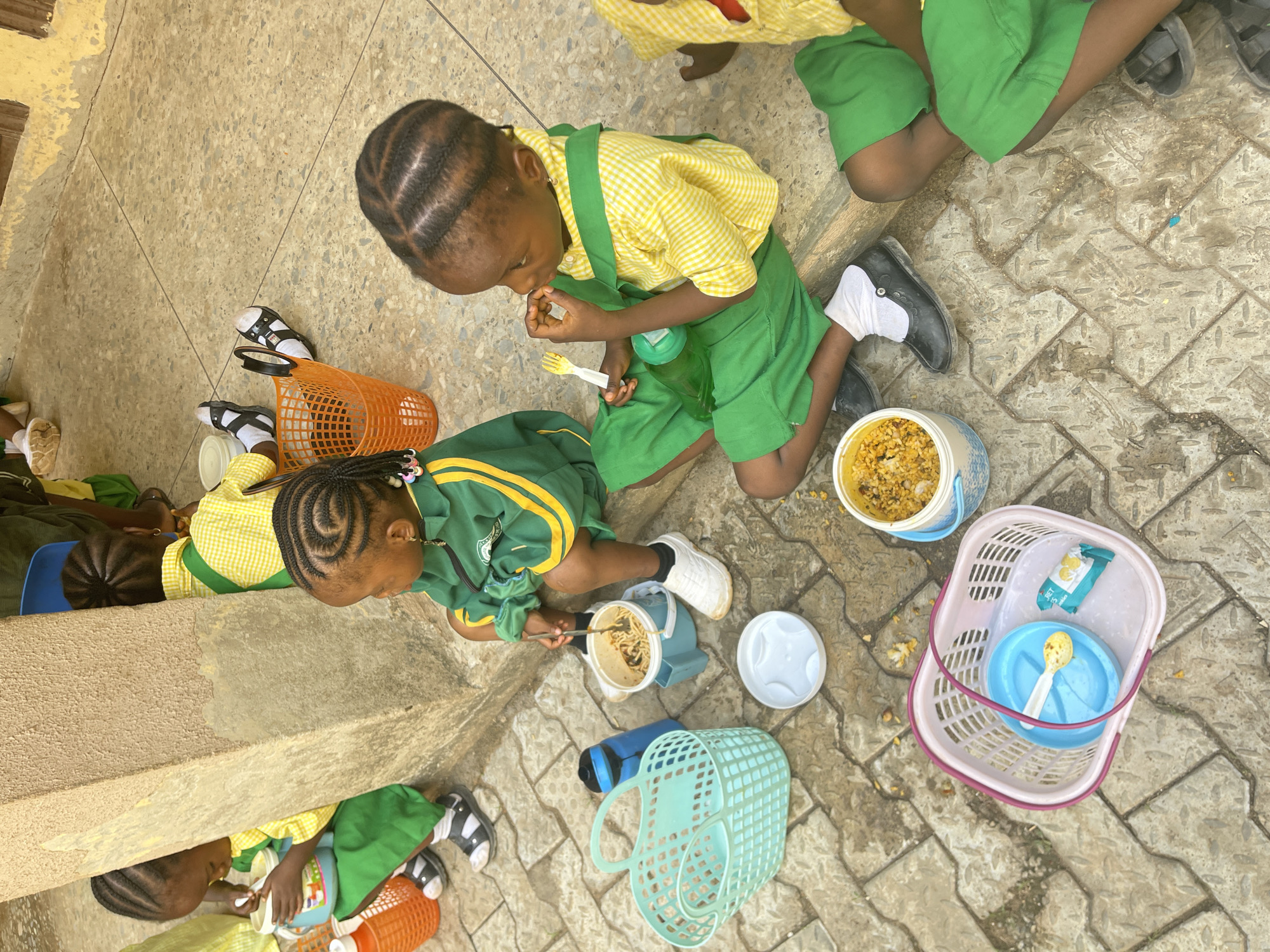 The image depicts a group of young children in school uniforms eating lunch outdoors in what appears to be Akwanga, Nigeria. The uniforms are primarily green and yellow. The children are sitting or kneeling on a tiled or paved surface. They are using plastic containers, plates, and utensils to eat. There are various plastic baskets scattered around, some containing food items or utensils. The setting appears to be a schoolyard or patio area.
