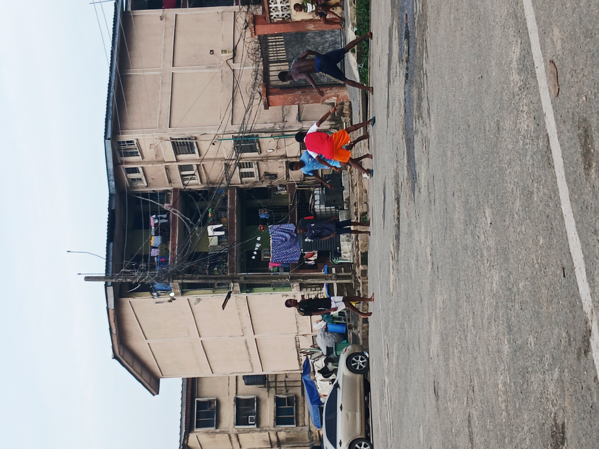 The image shows a street scene in a densely populated urban area likely in Lagos, Nigeria. A group of young men are seen in motion, possibly running or playing, on a concrete road. One man is shirtless, wearing dark shorts, while others are wearing various athletic attire in red, blue, and orange. A multistory building with visible wiring and laundry hanging from balconies forms the backdrop, indicating a residential area. A white car is parked near the building. The road has a white line marking, possibly indicating a lane division. The sky is overcast.