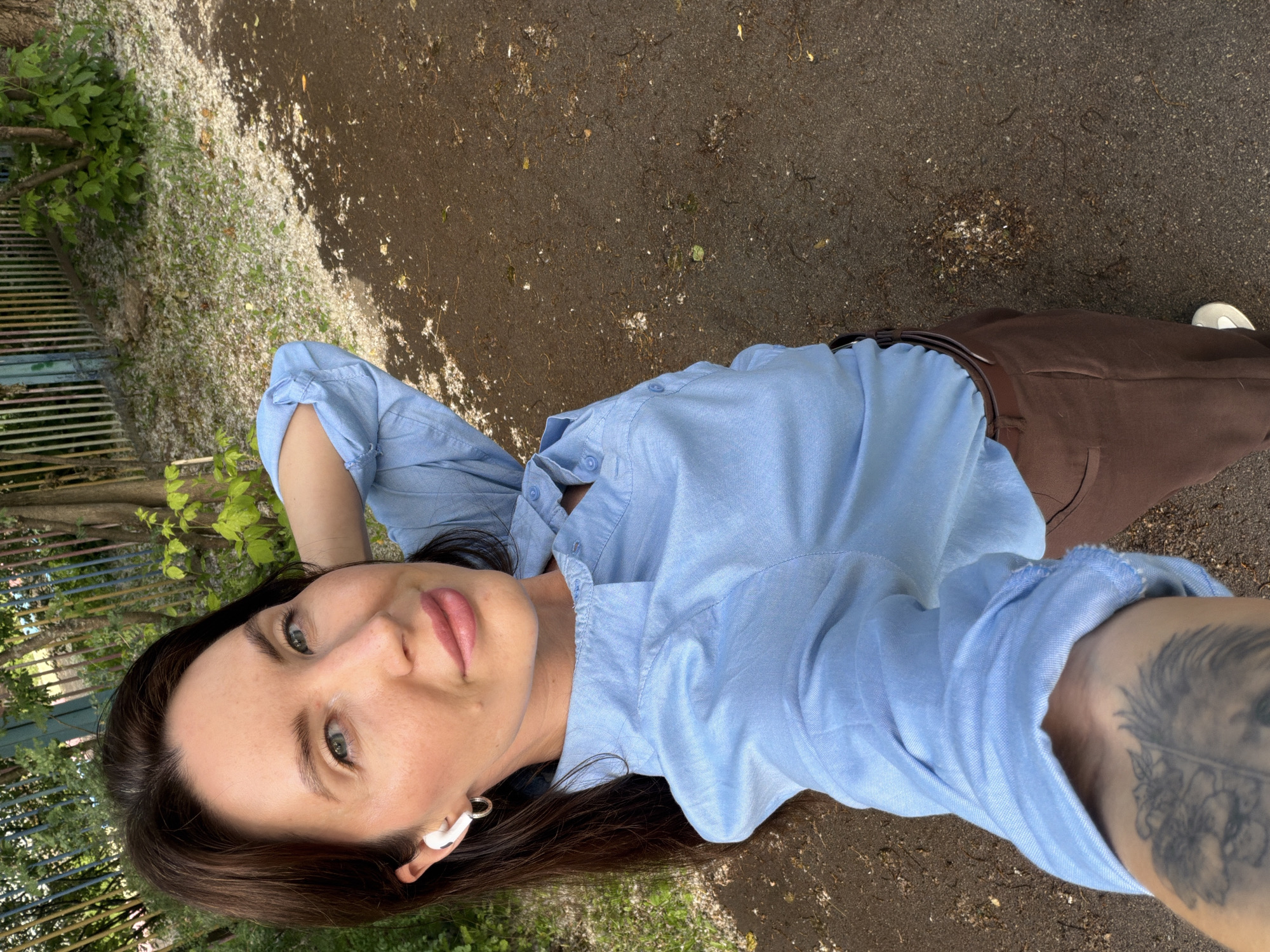 The image shows a woman with dark hair and blue eyes, wearing a light blue button-down shirt and brown pants. She has white earbuds in her ears and a silver hoop earring. Her right arm is behind her head. She is standing outdoors on a path, with trees and a fence visible in the background. The ground is covered with small white debris. The image appears to be taken in Perm, Russia.