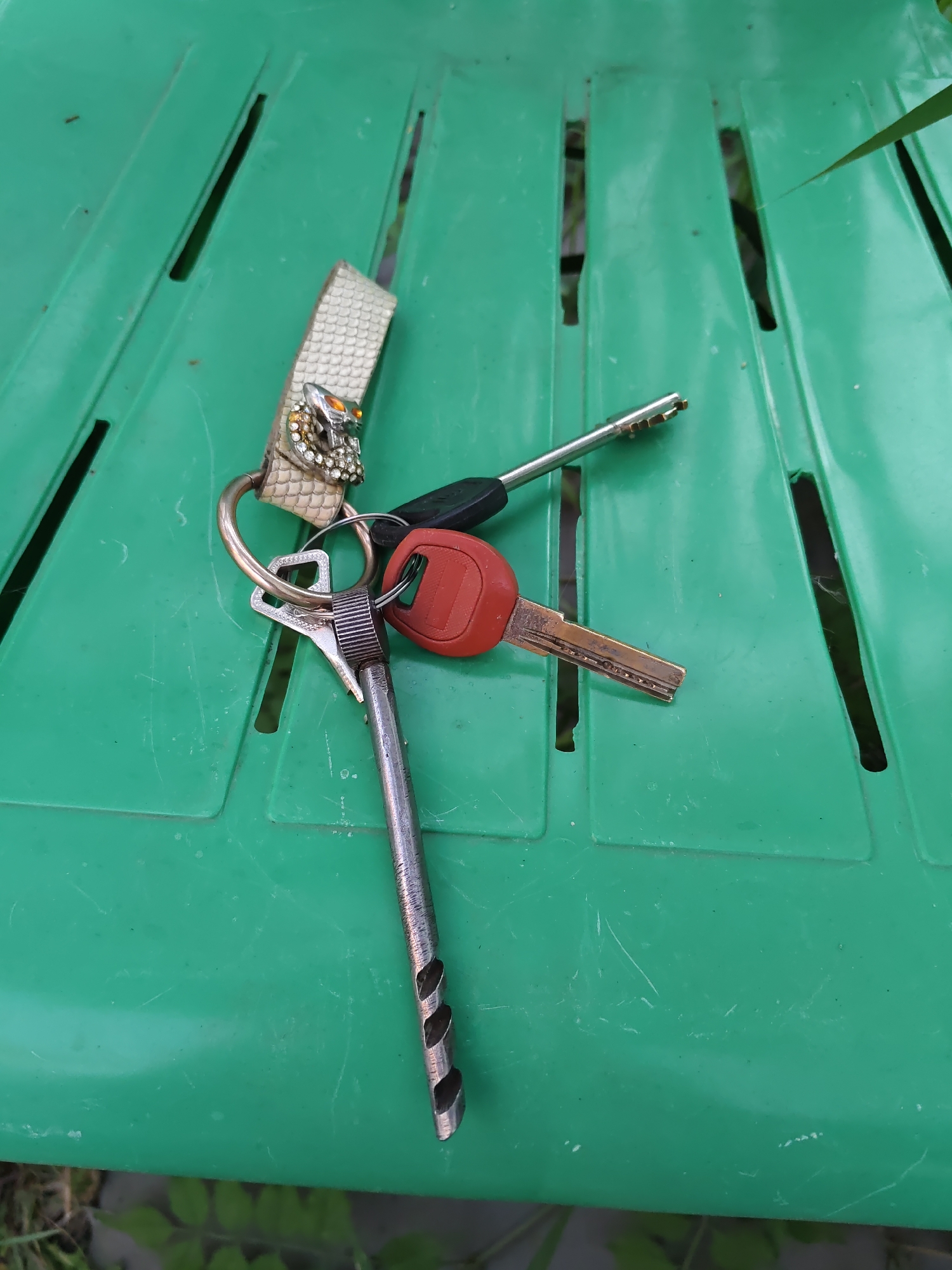 A set of keys with a decorative keychain, possibly a snake design with rhinestones and orange eye accents, hangs from a ring. The keys are placed on a green, slatted surface, likely a plastic chair. One key has a red plastic head, another has a silver metallic head and a dark tip, and a third key is fully metallic with a unique grooved shaft. The background is out of focus, showing hints of green foliage and a gray concrete-like surface. The overall impression is of everyday objects casually placed outdoors. The location is specified as Rostov-on-Don, Russia.