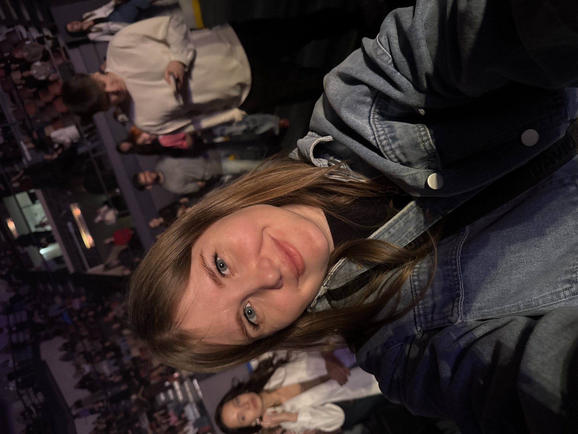 The image shows a young woman in a blue denim jacket taking a selfie. She has fair skin, light brown hair, and blue eyes. Behind her, out of focus, are several people, suggesting a crowded indoor space with many people. The image appears to be taken indoors, possibly at an event or gathering. Without more visual cues, it is not possible to confirm the location is in Perm, Russia.