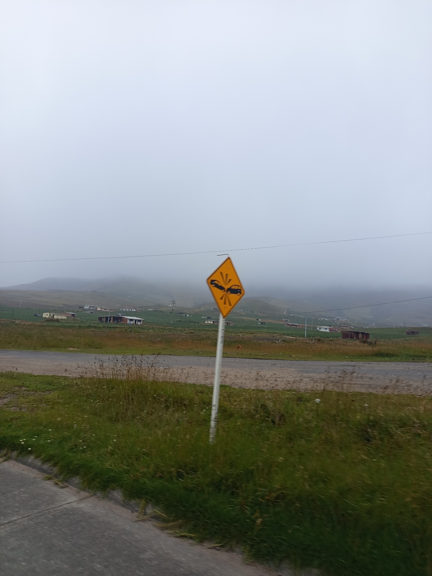 A yellow diamond-shaped warning sign with a black symbol depicting two cars colliding is positioned on a white pole in the foreground of the image. The sign is located on the side of a paved road that curves into the distance. To the left of the road is a concrete sidewalk. The background features a rural landscape with rolling hills covered in green grass and scattered houses. The sky is overcast and foggy, obscuring the tops of the hills. The scene appears to be in a sparsely populated area with a muted color palette due to the weather conditions. There are no people or animals visible in the image. The image was taken in the city of Tona, Colombia.