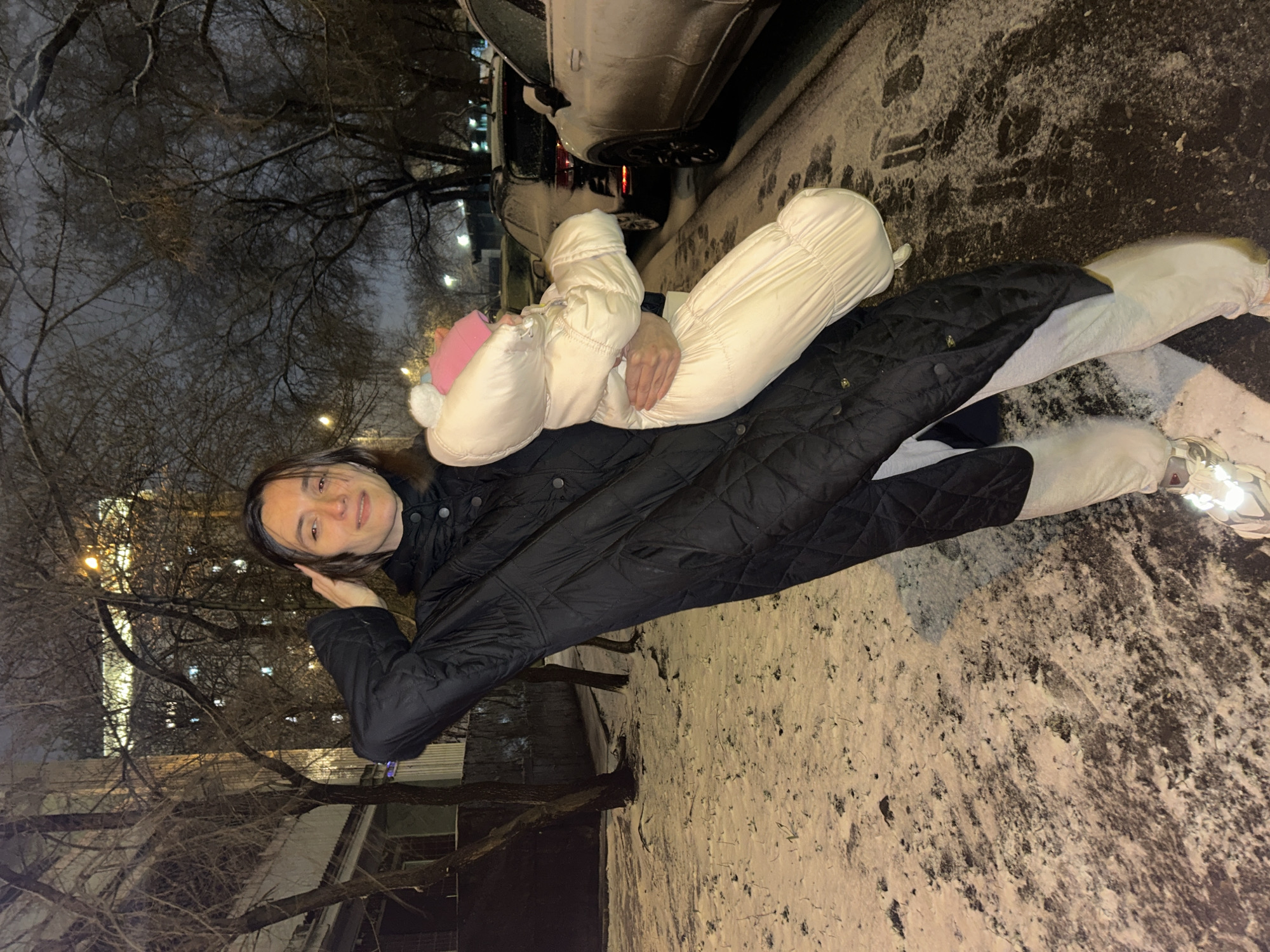 A woman is outdoors at night in winter in Moscow, Russia. She is lying on a snowy surface, possibly a sidewalk, and holding a young child, who is bundled in a white snowsuit with a pink hat. The woman is wearing a black quilted jacket and white pants. A car is parked behind her on the snowy street. Bare tree branches are visible against the night sky, with building lights in the background. Snow is visible on the ground and possibly the car.