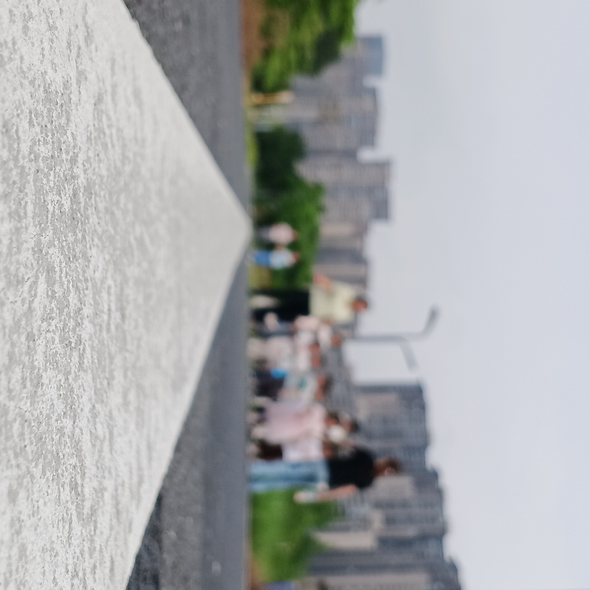 The image, originally oriented sideways, depicts an outdoor scene with a strong shallow depth of field. In the immediate foreground, a sharply focused, textured white line runs diagonally across the frame, suggesting a painted curb, pathway marking, or barrier. Its surface shows a rough, granular quality.
To the right of this dominant foreground element, the scene becomes progressively blurred. In the midground, several indistinct figures of people are visible, some appearing to be walking or standing on a path. Their forms are too blurry to discern individual features, clothing details, or emotions, but they suggest a social gathering or casual movement. Patches of green foliage are also present in this area.
The background is heavily blurred, revealing an urban or suburban landscape dominated by numerous tall, multi-story buildings, characteristic of residential or commercial architecture found in cities like those in Shunde District, China. The sky above is bright and overcast, indicating a daytime setting without direct sunlight. No specific activities other than the implied movement of people are clearly discernible. There is no visible text in the image.