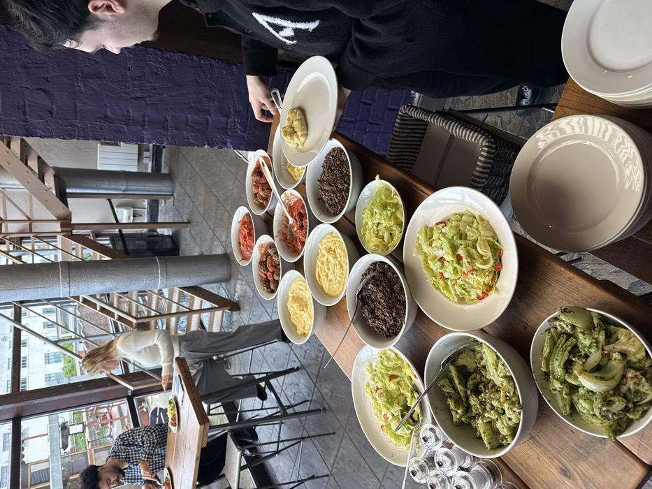 The image depicts an interior scene within a modern restaurant or cafe in London, United Kingdom, likely situated within a repurposed industrial building, characterized by large windows and an exposed metal and wood staircase.
In the foreground, a male individual, viewed from the right in profile, wearing a dark jacket with a white 'A' logo, is serving himself from an extensive buffet. He holds a white plate and is scooping a yellowish, mashed food item from one of numerous white ceramic bowls arranged on a long wooden table. The buffet presents various dishes in white bowls: multiple servings of meatballs in red sauce, plain red sauce, several bowls of the yellow mashed food (possibly polenta or mashed potatoes), dark granular food (such as lentils or minced meat), chopped green salads with red flecks, and green roasted vegetables in a pesto-like sauce. A stack of clean white plates is visible to the right of the buffet line.
In the midground, to the left, two individuals are seated at a high wooden table with metal legs. A male with dark curly hair, wearing a plaid shirt, is actively eating from a plate. Opposite him, a female with blonde hair, dressed in a white fluffy sweater and grey trousers, is seated and engaged with a digital device (tablet or phone) on the table.
The background features large windows offering a view of an exterior grey building with multiple windows, characteristic of an urban environment. An industrial-style metal staircase with wooden treads ascends on the left side, and a purple-painted brick wall is visible on the right.