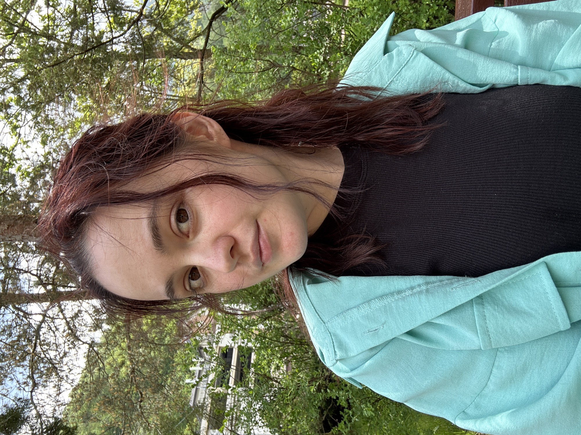 A woman with dark auburn hair is pictured in a slightly off-center medium shot, wearing a black top and a light turquoise button-up jacket. The background appears to be a wooded area with green foliage and trees. There is a glimpse of a building or structure with white walls visible through the trees. Based on the presence of deciduous trees and architectural style, it's highly likely to be Estosadok, Russia, given the context provided in the query. The subject is looking directly at the camera with a slight smile. The image is taken outdoors during daylight hours.