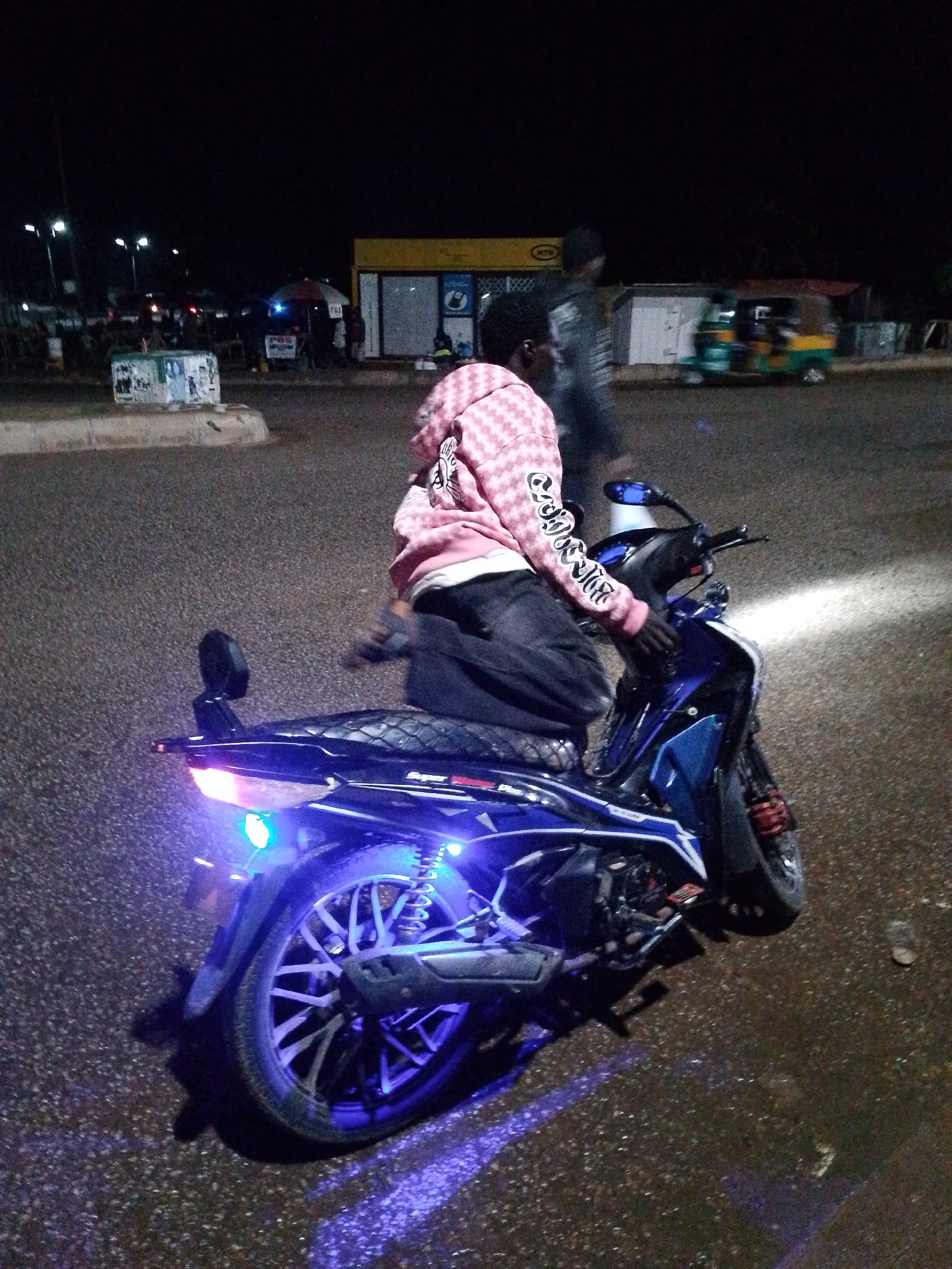 The image captures a nighttime scene on a paved road in Bukuru, Nigeria. A young Black male, wearing a pink checkered hooded sweatshirt with white lettering on the sleeves and dark pants, is seated on a blue and black motorcycle. The motorcycle features illuminated blue lights on its rear and sides, as well as a bright white headlight. In the background, there are several structures, including a yellow building with windows and signage, a white building, and what appears to be a market stall with an umbrella. A multi-colored three-wheeled vehicle (likely an auto-rickshaw) is visible in the mid-ground. The ground is wet, suggesting recent rain, and reflections of the motorcycle's lights are visible on the surface. Other individuals are present in the background, some blurred due to motion. The overall atmosphere is that of a busy urban setting at night.