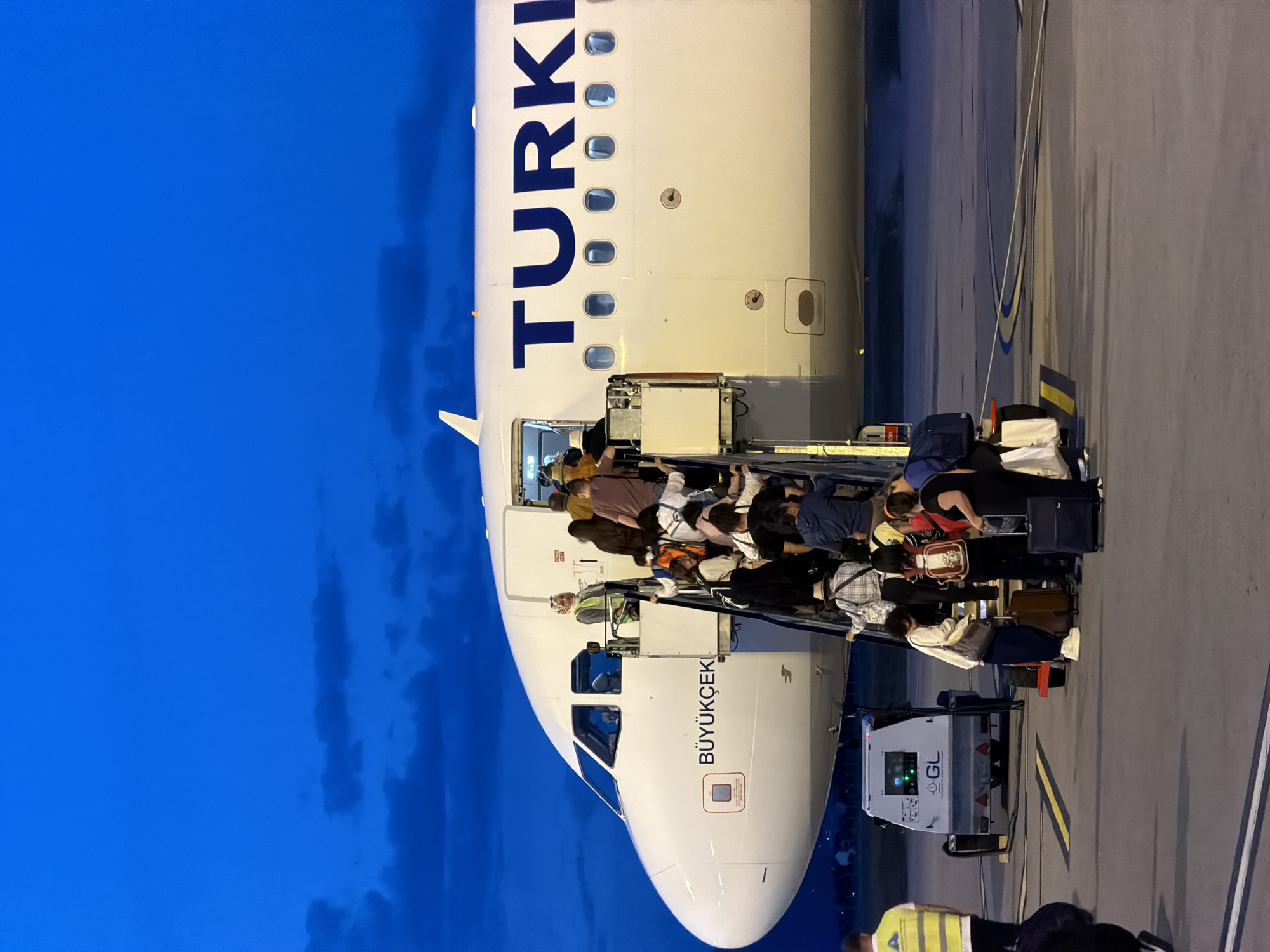 The image shows passengers disembarking a Turkish Airlines airplane via stairs to the tarmac. The plane's front portion, with the word "TURKI" visible, is on the left side. Passengers with luggage are descending the stairs. The nose of the plane has the inscription "BÜYÜKÇEK". A "GL" labelled vehicle is parked under the stairs. The scene appears to be at an airport at dusk, under a dark blue sky. The location is Podgorica, Montenegro, based on the airline serving that city.
