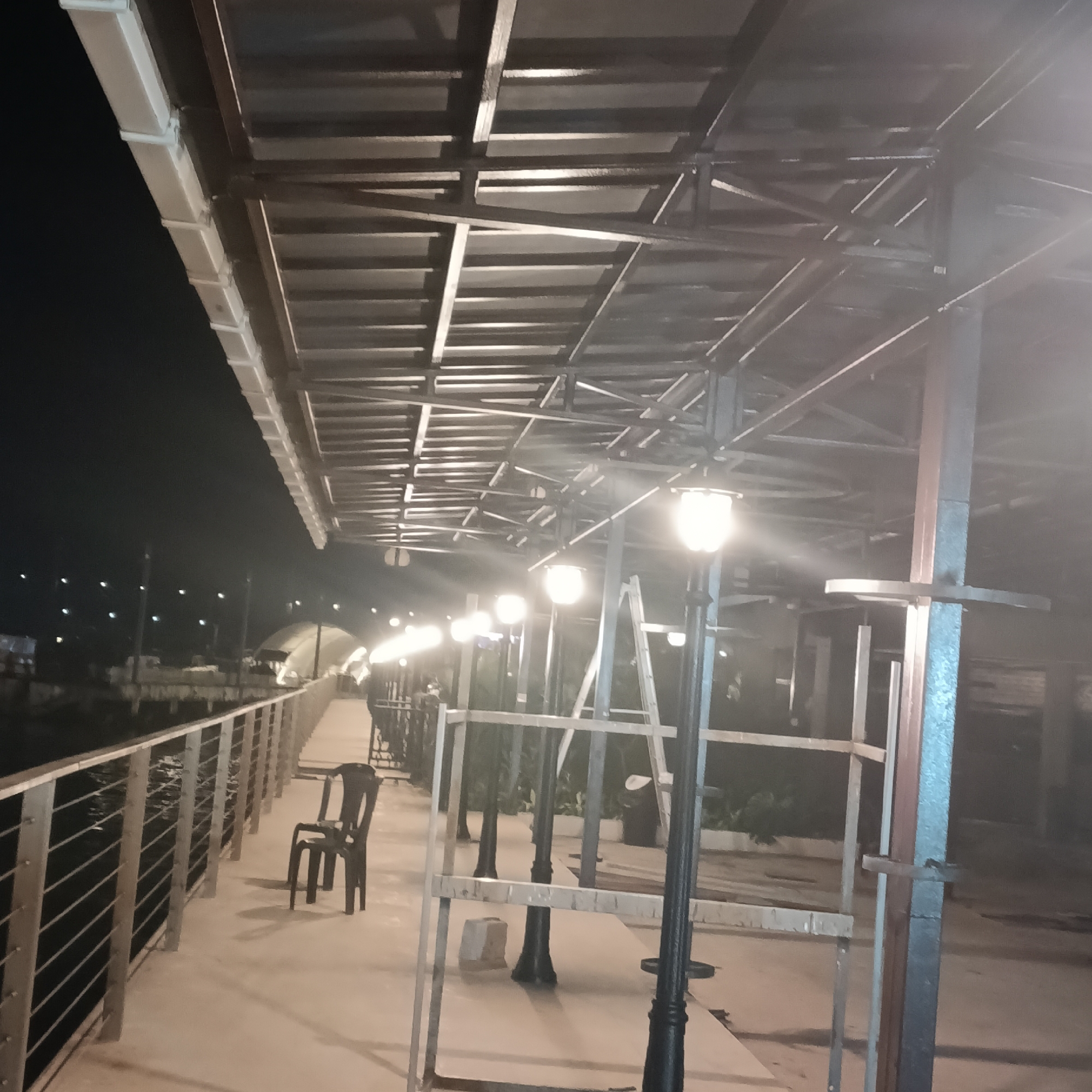 The image depicts an outdoor walkway in Panaji, India, likely at night, illuminated by several streetlights. The walkway is lined with a metal railing on one side. A chair is positioned alongside the railing. Above the walkway, a metal structure forms a partial roof. Scaffolding is erected in the walkway.