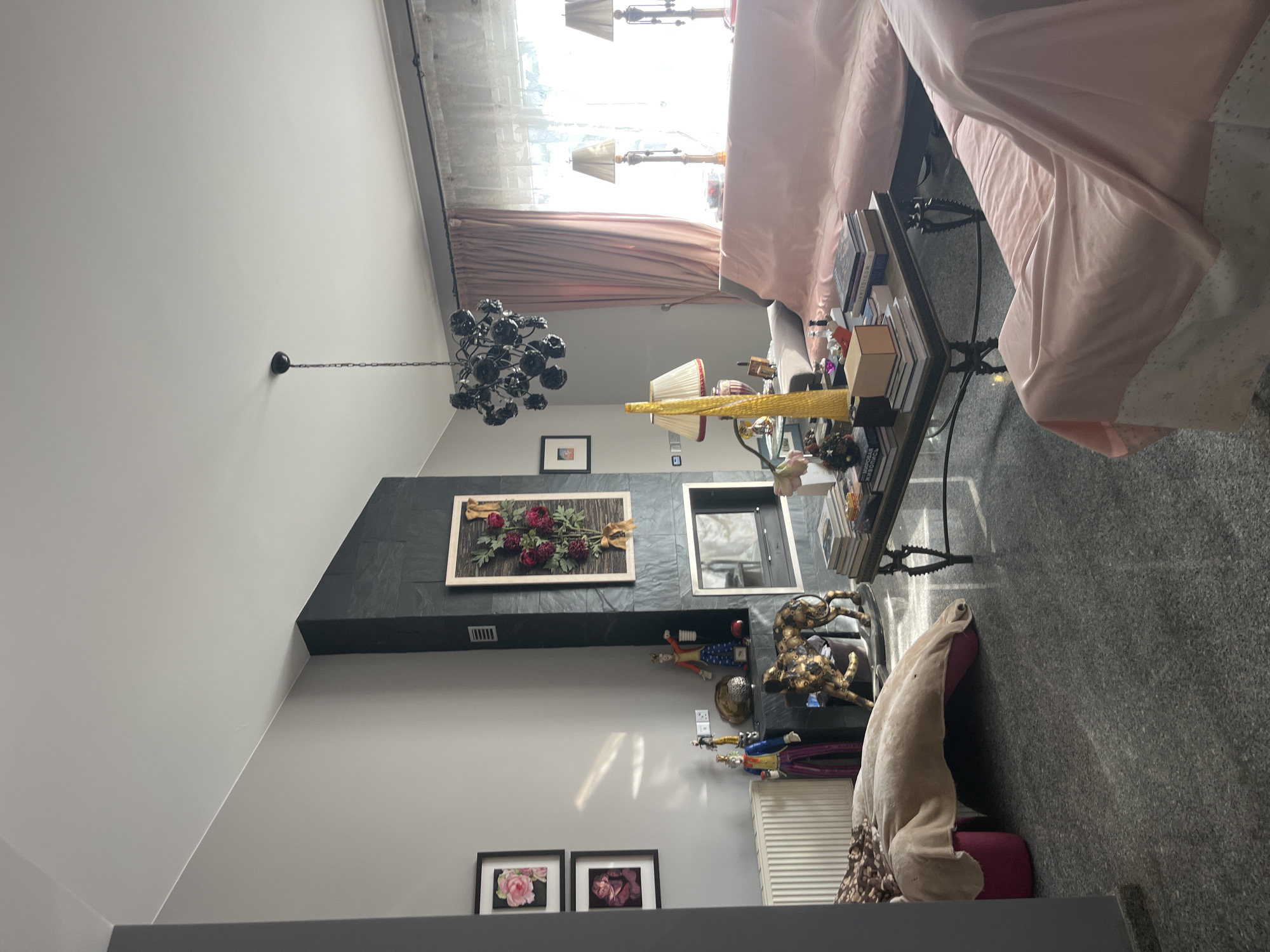 The image depicts an indoor living room setting, likely in Parekklisia Community, Cyprus. The room features a dark stone fireplace on the left wall, adorned with a framed floral arrangement. To the left of the fireplace, two framed floral pictures hang on a light gray wall. In front of the fireplace, there are two decorative figurines, one in a colorful outfit and another in a suit, standing next to a golden leopard statue. A radiator is visible on the far left wall.
In the center of the room, a dark coffee table is laden with books and decorative items. To the right, two long sofas, draped in light pink fabric, are arranged facing the coffee table and a large window. Two table lamps with beige shades are placed on either side of the window, which is covered by sheer white curtains and heavier pink drapes. A modern black chandelier hangs from the ceiling above the sofas. The floor is a dark, speckled tile. The overall impression is of a well-furnished, somewhat eclectic living space.