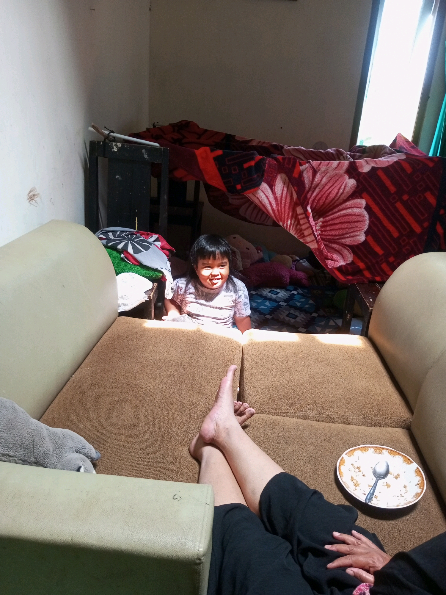 The image captures a child sitting behind a couch in a dimly lit room. The child is smiling and looking towards the camera. A blanket with a floral pattern is draped over a piece of furniture in the background, creating a makeshift tent-like structure. The room appears to be a living space, possibly in a residential area in Krasak, Indonesia, given the style of furniture and the overall ambiance. A dirty plate with a spoon rests on the couch in the foreground, suggesting a meal has recently been consumed. The scene is illuminated by sunlight streaming in from a window, casting shadows across the room.