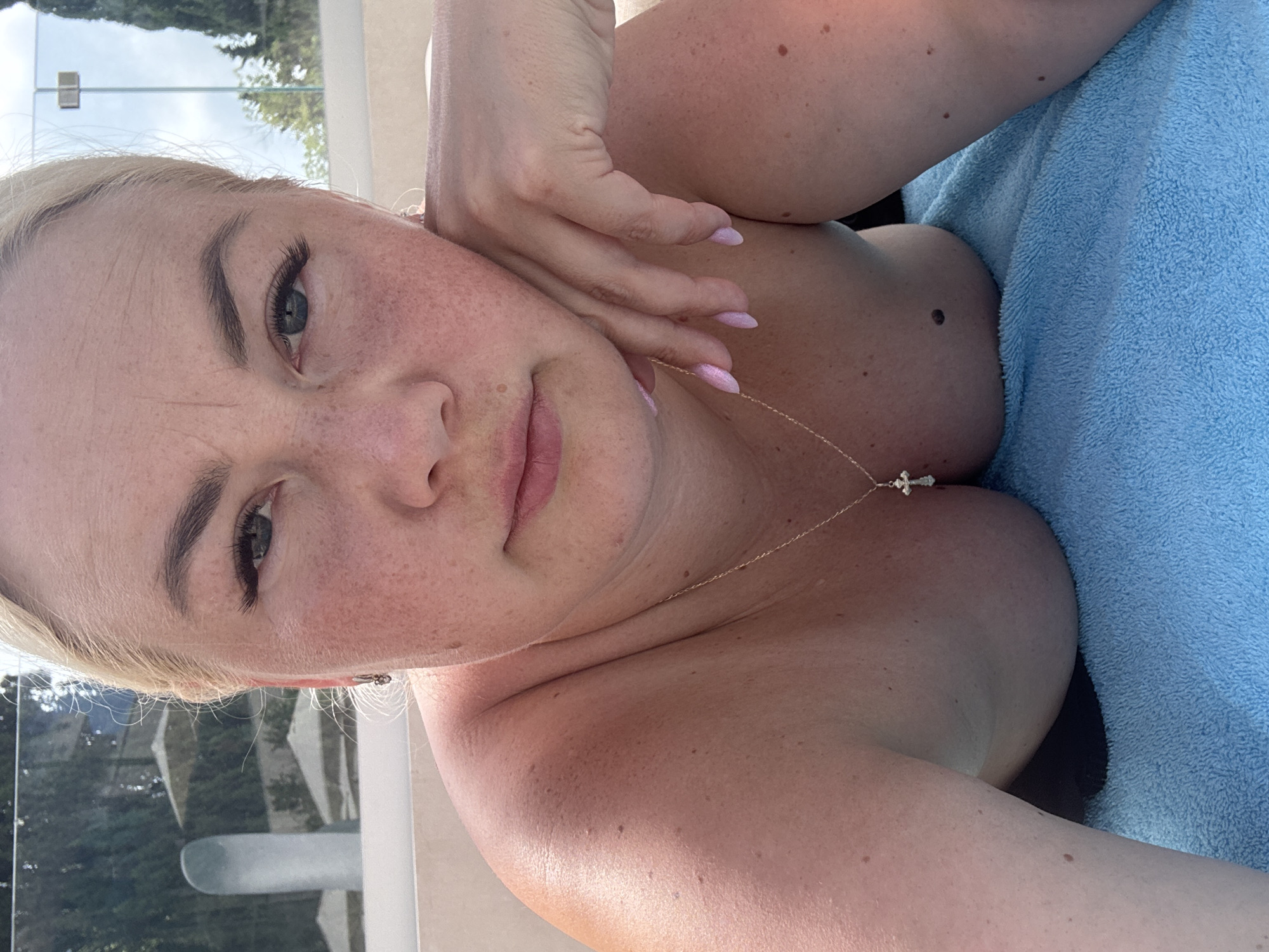 A woman is lying down outdoors on a blue towel. She has fair skin with freckles and blonde hair. She is wearing a necklace with a cross pendant. Her eyes are open and looking at the camera. Her right arm is bent and her hand is resting on her cheek. The background appears to be a poolside area with trees and lounge chairs.