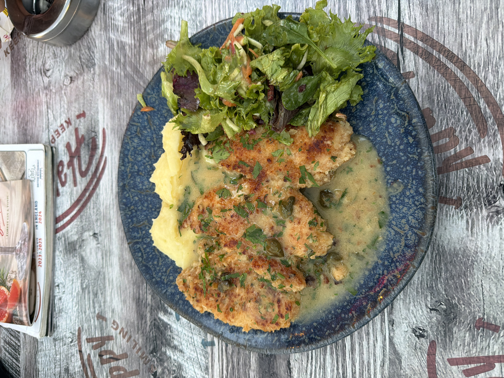 A close-up, overhead shot shows a plate of food on a weathered wooden table. The meal consists of mashed potatoes, two breaded chicken cutlets in a creamy sauce with capers and parsley, and a side salad of mixed greens. The plate is dark blue with a textured rim. To the left of the plate is a stack of magazines or brochures. The top one has a cover with an illustration of fruit and text that includes "FOREST GIN & TONIC" and "IN ENGLAND 2023 & 2024". The table surface has faint reddish-brown lettering, possibly from a placemat or branding, with partial words visible such as "KEEP", "etty", and "UMPHING". The scene appears to be outdoors, suggested by the natural light and the rustic table. The context provided indicates the location is Burgas, Bulgaria, though no explicit visual cues in the image confirm this. There is no indication of the time of day or weather. No people are visible in the frame. The overall impression is a casual meal at a restaurant or outdoor cafe.