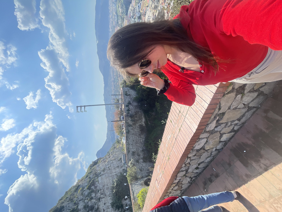 The image depicts an elevated panoramic view of the city of Meta, Italy, on a clear, sunny day with a bright blue sky and scattered white cumulus clouds.
In the foreground, a female individual, appearing to be in her twenties or thirties with long brown hair, is positioned with her right profile visible. She is wearing a red long-sleeved top, light-colored trousers, and round-framed sunglasses, which she is adjusting with her right hand. A black scrunchie is on her left wrist. To her left, partially visible, is another individual from the torso down, wearing a dark blue puffer vest over a red garment, light blue jeans, and dark shoes, walking away from the camera. Both subjects stand on a terra cotta tiled walkway bordered by a stone wall topped with red bricks, which serves as a railing. Sunlight casts distinct shadows on the walkway.
The midground features a winding road traversing the side of a steep, rocky mountain on the left. The road is supported by a stone retaining wall, and several vehicles are visible on it, including cars and a van. Road signs with white arrows on a black background are present, indicating curves. Beneath the road, an arched stone structure is visible. A tall, light-colored utility pole with three insulator assemblies stands prominently against the sky. Sparse vegetation, including trees and bushes, dot the landscape between the road and the foreground.
In the background, the city of Meta stretches across the landscape, characterized by numerous light-colored buildings with reddish roofs densely packed together. Distant mountains frame the horizon beyond the cityscape.