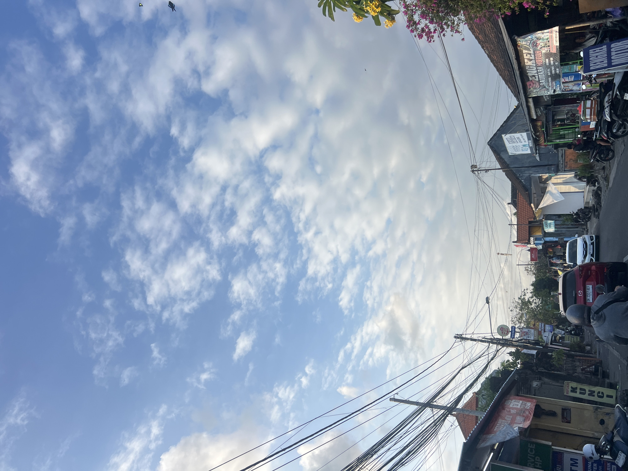 The image depicts a street scene in Denpasar, Indonesia, under a partly cloudy sky. The street is lined with buildings, some of which appear to be small businesses or residences, with traditional tiled roofs. Overhead, a dense network of electrical wires stretches across the frame, a common sight in many Indonesian cities.
In the foreground, several motorbikes are parked along the side of the road. A red car is in motion in the center of the street, and a white car is visible further down. A person wearing a grey hoodie and helmet is riding a motorbike, their back to the camera. Another person is standing in front of a shop on the right side of the street.
Several signs are visible on the buildings. One sign on the right reads "BENGKEL SAUDARA MOTOR," indicating a motorcycle repair shop. Other signs are in Indonesian, with some partially obscured or unreadable. Yellow and pink flowers are blooming on a tree on the right side of the image, adding a touch of color to the urban landscape. The overall impression is of a bustling, everyday street in a tropical city.