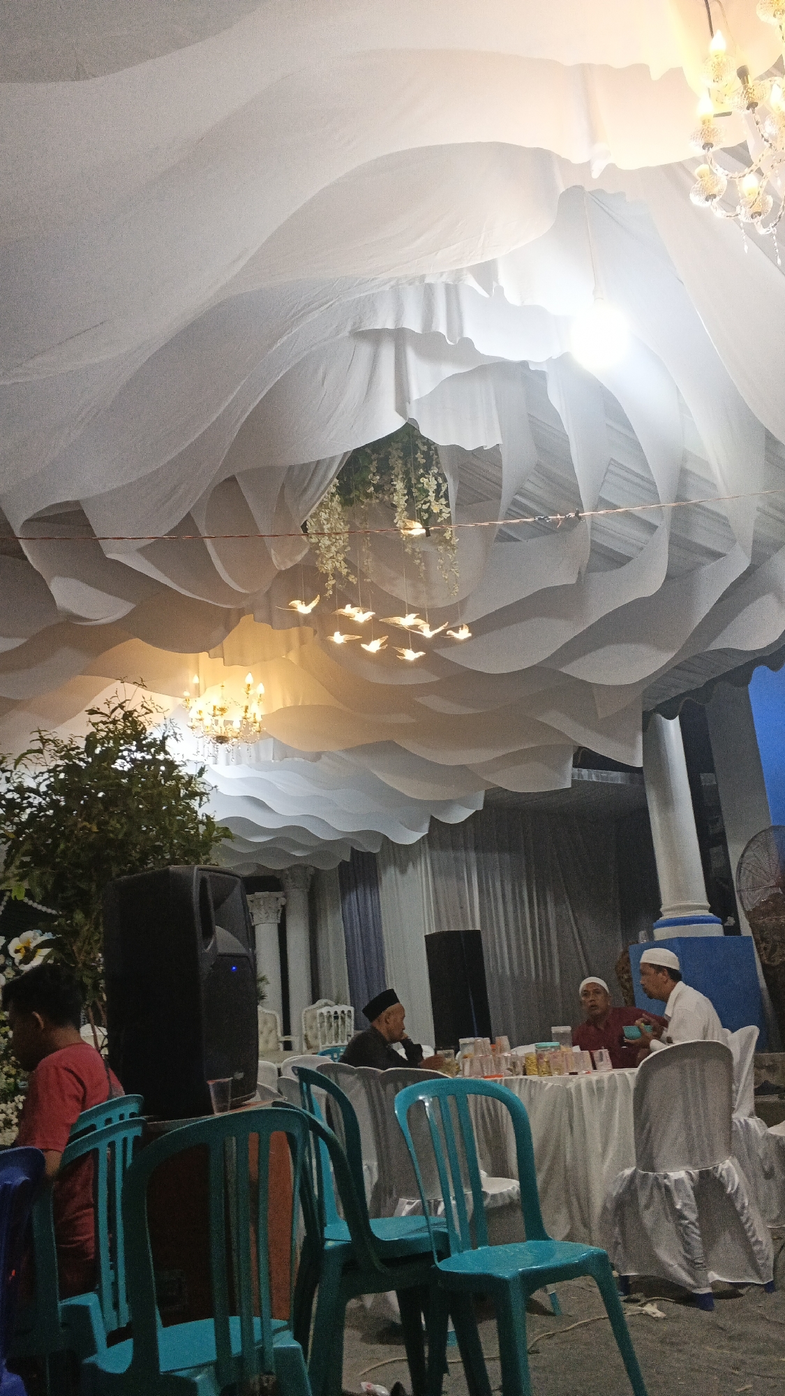 The image depicts an indoor gathering space, likely a venue for an event, decorated with white flowing fabric draped from the ceiling, creating a wavy effect. Chandeliers and hanging bird-shaped lights are visible, along with floral decorations suspended from the ceiling. Several people are present, some seated at tables covered with white tablecloths. One group of men, dressed in traditional Islamic attire, are seated at a table, appearing to be in conversation. The environment suggests a formal or semi-formal event. The setting is identified as Soko, Indonesia.