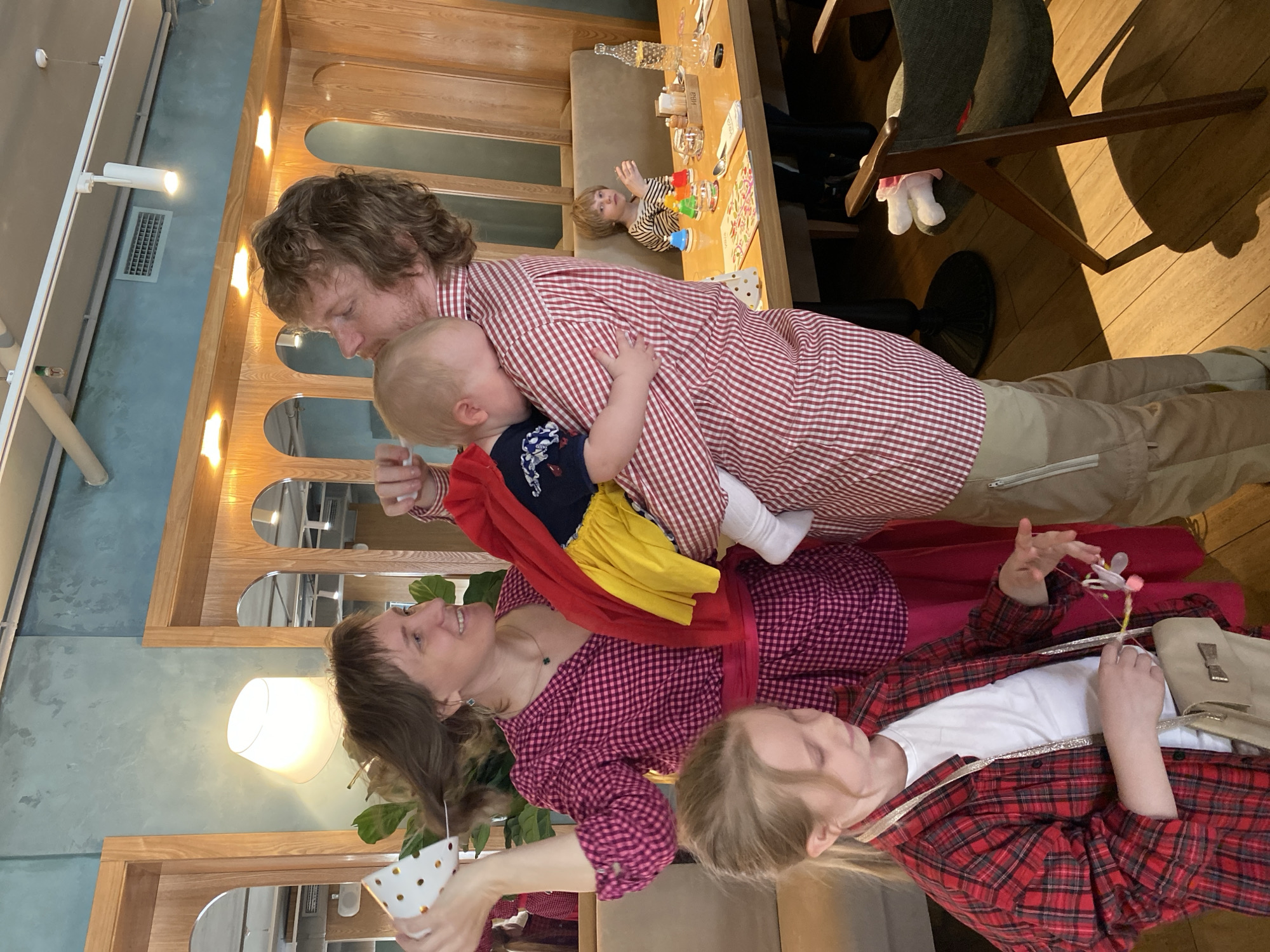 The image shows a family of four in what appears to be a restaurant. A man with curly hair is holding a baby in his arms, who is dressed in a navy blue shirt with a yellow skirt and red cape. A woman in a pink and purple dress is smiling and looking at the baby. A young girl in a red and black checkered shirt is next to the woman, holding a wand.
The restaurant has wooden tables and chairs. There is another child sitting at the table. The walls are painted a light blue color with wooden accents. Based on the location metadata, this scene may be in Yekaterinburg, Russia.