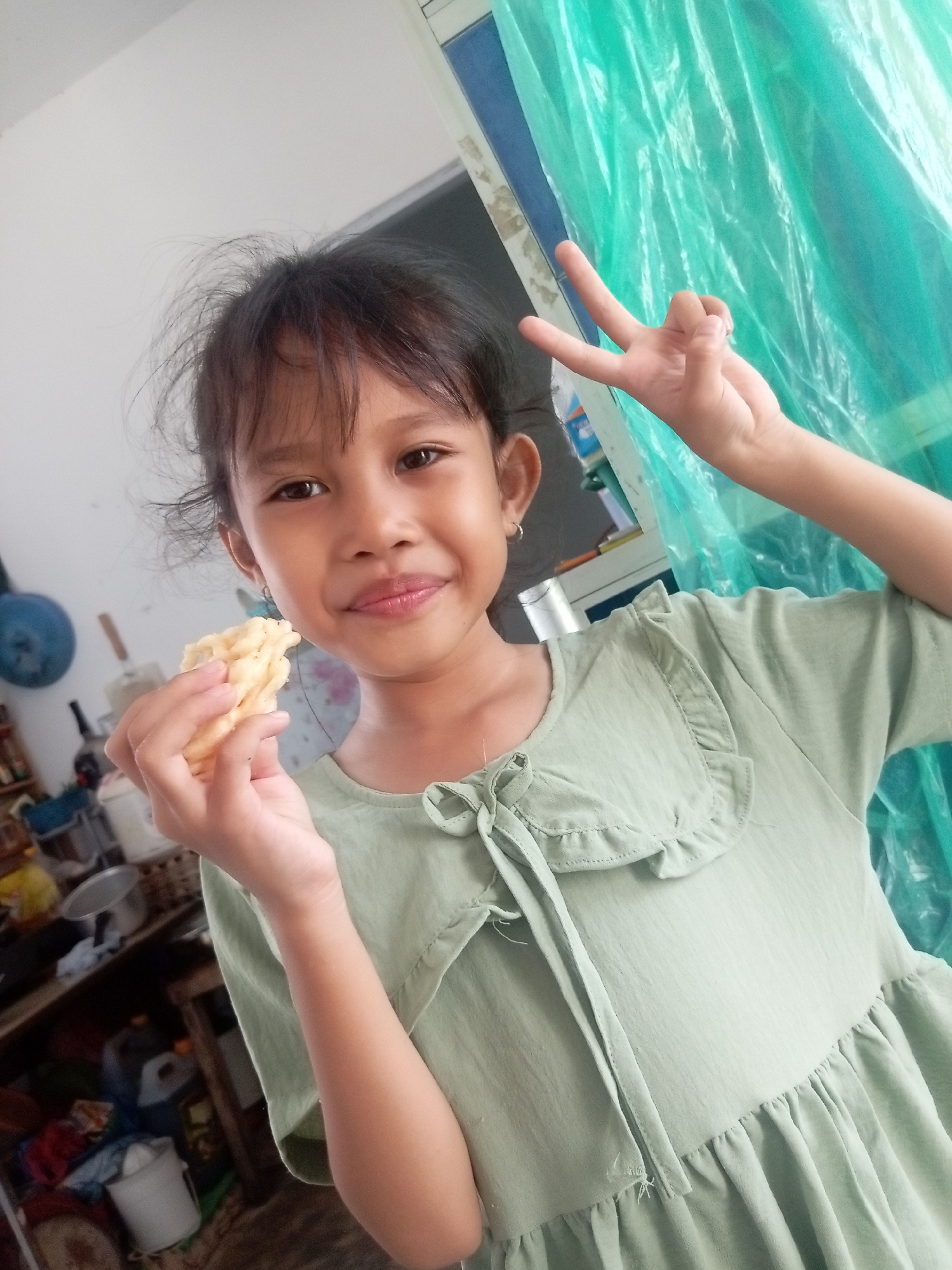 A young girl, with dark hair and brown eyes, is smiling at the camera while holding a light-colored snack in her left hand. She is wearing a light green dress with ruffled details around the neckline and a tie in the front. Her right hand is raised, forming a peace sign. The background is blurred but appears to be an indoor setting with shelves filled with various items, including containers and clothing. A bright green plastic sheet hangs on the right side of the frame. The overall lighting suggests it is daytime. The image appears to be a casual, personal photograph.