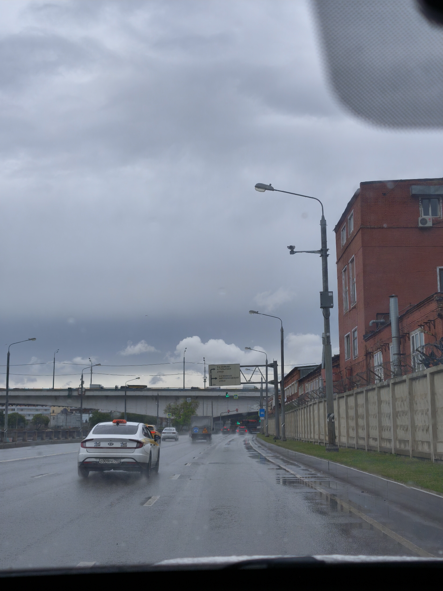 Here is a detailed description of the image:
**Scene:** A street scene on a wet, cloudy day in Moscow, Russia. The image appears to be taken from inside a vehicle, looking out through the windshield.
**Environment:** Overcast sky with heavy clouds indicating rain. The road surface is wet, with puddles and reflections of the surrounding environment.
**Objects:**
* **Vehicles:** Several cars are visible on the road. A taxi with a visible license plate "E009PK 750" is in the foreground. Other vehicles include cars, trucks, and possibly buses further down the road.
* **Buildings:** A large brick building is on the right side of the road, behind a tall concrete fence with barbed wire. The building appears industrial or commercial.
* **Infrastructure:** A large overpass/bridge is visible in the background, supported by concrete pillars. Streetlights and signage are also present.
* **Signage:** Street signs written in Russian direct traffic to various routes (e.g., 3-е кольцо Старообрядческая улица, Автозаводская улица).
* **Greenery:** Patches of green grass visible along the roadside and beyond the fence.
**Actions/Interactions:**
* Vehicles are driving on the road.
* Rain is falling, impacting visibility and creating reflections on the road.
**Location:**
Based on the signage and architectural style, the scene is likely in an industrial or commercial area of Moscow, Russia, near the 3rd Transport Ring Road and various streets named in Russian (e.g., Старообрядческая улица, Автозаводская улица).