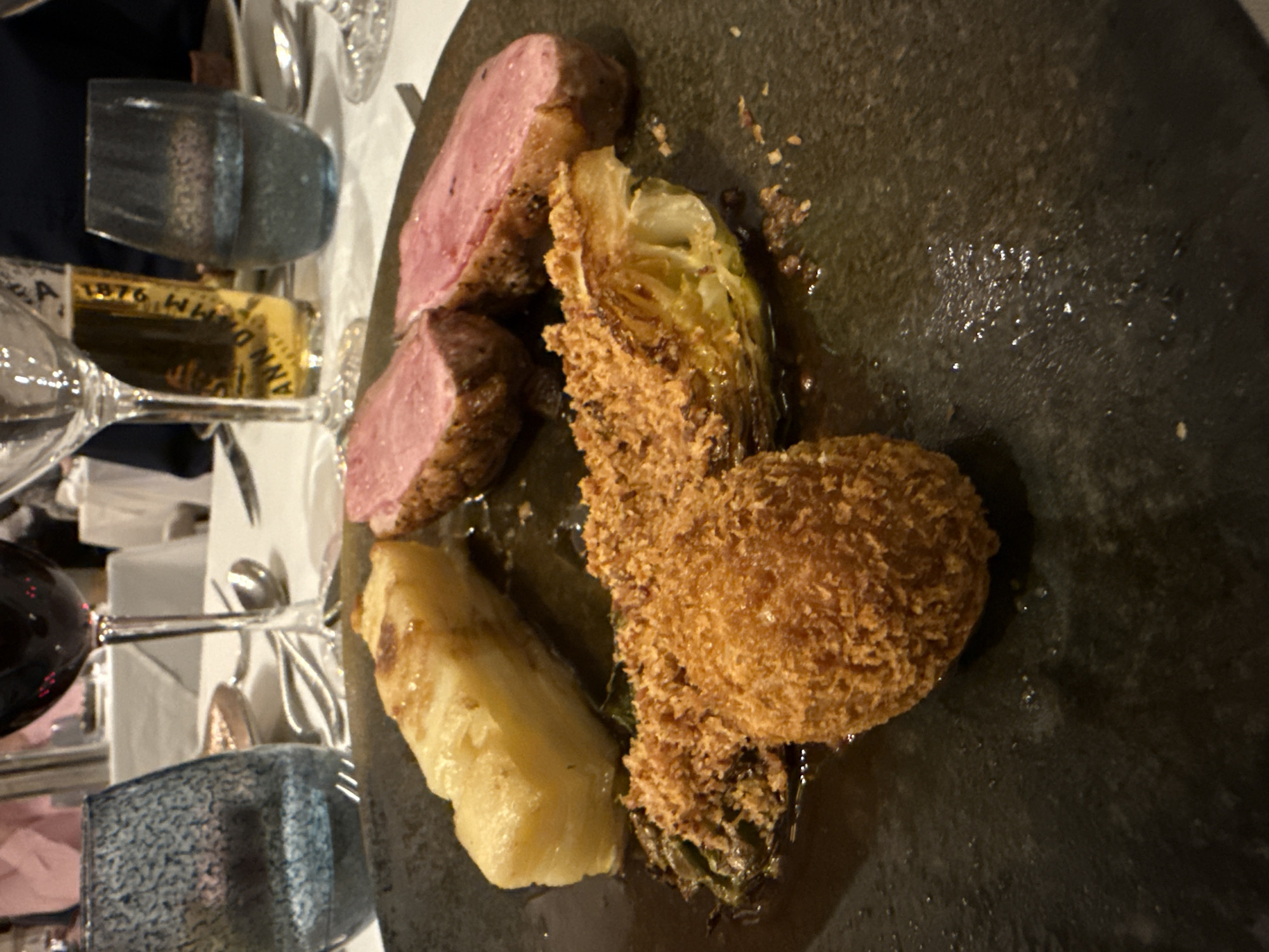 The image captures a plate of food, seemingly taken at a restaurant. The primary elements are slices of what appears to be rare duck or lamb, accompanied by fried, breaded Brussels sprouts, and a soft white vegetable. The plating is rustic, on a dark grey or black textured plate. In the background, the dining table setting is visible, including wine glasses, water glasses, cutlery, and a bottle of alcohol, likely wine or whisky. Due to the presence of a bottle labeled "1876" it might be related to a special occasion. The scene is indoors, likely a restaurant. Without further context, the location is unknown, and I can not relate to Glenridding, United Kingdom.