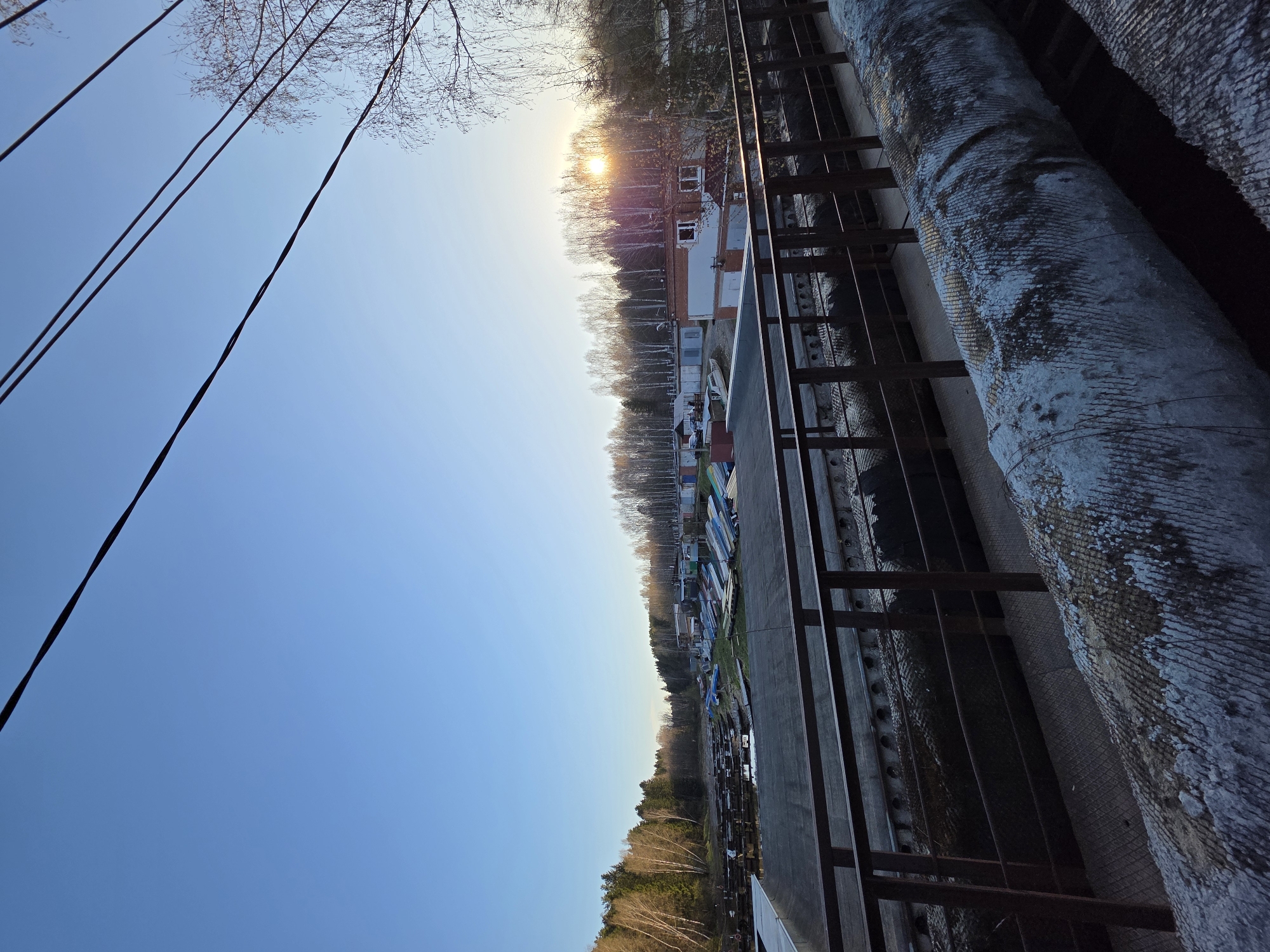The image is taken from an elevated position, possibly a building rooftop, in Chaikovsky, Russia, facing a landscape at sunrise or sunset. The foreground contains industrial elements: insulated pipes, a metal railing, and wiring extending into the clear blue sky. The midground reveals a riverbank lined with boats and other watercraft stored neatly alongside a narrow road. Beyond, there is a row of buildings, some with windows visible, partially obscured by a line of trees. The background showcases a thick forest and the bright sun illuminating the scene through the trees. The overall atmosphere suggests a blend of urban and natural environments in the setting of Chaikovsky, Russia.
