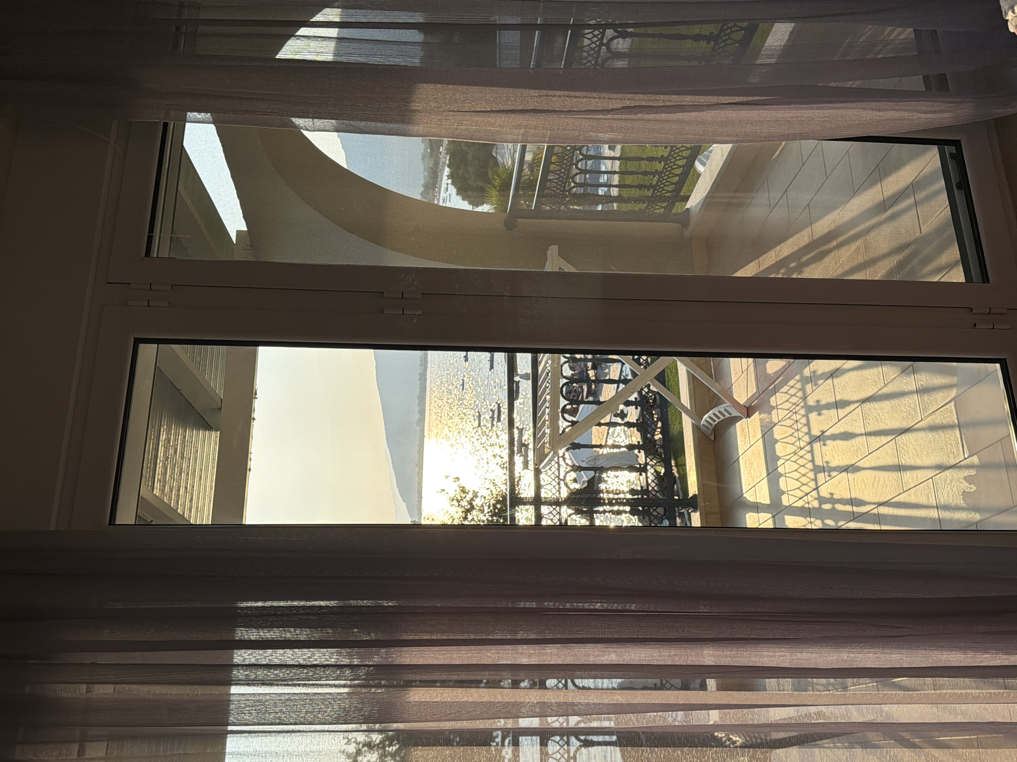 The image depicts a view from a balcony overlooking a body of water in the Municipal Unit of Alyzia, Greece, during what appears to be sunset. The foreground is dominated by sheer curtains, one on the left and one on the right, partially obscuring the view. Through the glass doors, a balcony with tiled flooring is visible, illuminated by warm, angled sunlight casting distinct shadows. Beyond the balcony railing, which features an ornate metal design, a body of water is seen, reflecting the golden light of the setting sun. Several small boats are dotted on the water. In the distance, a coastline with hills and trees is visible, creating a serene, picturesque landscape. The architecture suggests a resort or residential area with balconies and open arches.