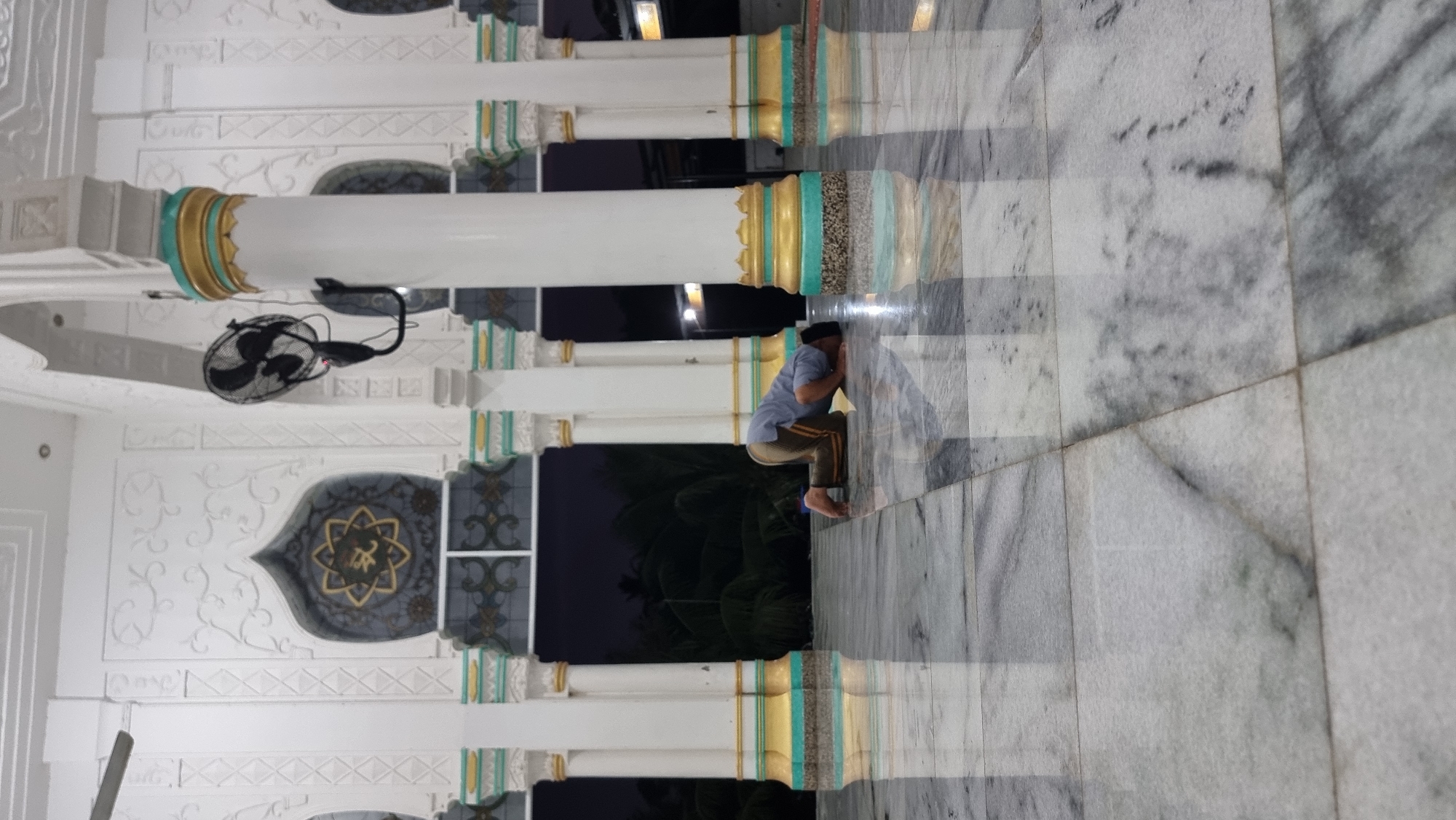 A man is praying on the marble floor of a mosque in Lapang Timu, Indonesia. He is wearing a light blue t-shirt and tan pants with orange stripes, and has a black skullcap on his head. The man is in the sujud position, with his forehead touching the floor. The mosque has white pillars with gold and teal accents, and intricate white decorative carvings on the walls. A black oscillating fan is mounted on the wall behind the man. Through the open archways, palm trees and a dark sky are visible. The marble floor is highly reflective, mirroring the pillars and the man.