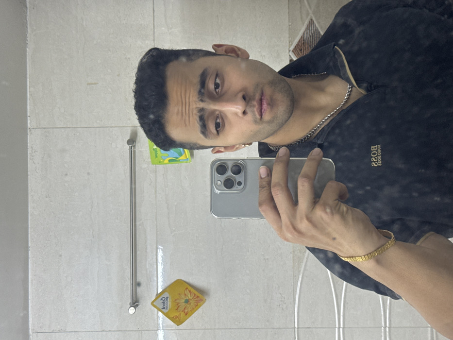 A male individual is captured in a mirror selfie in an indoor setting, identified as Kolkata, India. He is depicted from the chest up, with dark, styled hair, a slight stubble, and wearing a dark collared shirt with the "BOSS HUGO BOSS" logo visible in reverse on his left chest. Accessories include a silver-colored chain around his neck, a small gold-colored hoop earring in his left ear (viewer's right), and a gold-colored bracelet on his right wrist (viewer's left). He holds a light gray smartphone, featuring a prominent triple-lens camera system, in his right hand to photograph his reflection. The background, reflected in the mirror, shows a wall covered with light-colored rectangular tiles. A horizontal silver-colored metal towel rail is mounted on this tiled wall. To the left of the rail, a yellow, square-shaped item with a floral design is adhered, and to the right, a green, irregularly shaped item with a blue/green design is also affixed. The subject's expression is neutral with slightly furrowed brows and pursed lips.