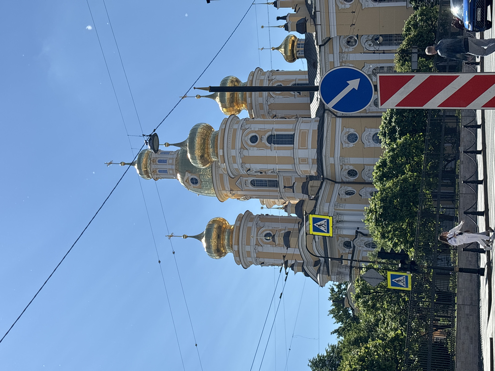 This is a medium shot photograph taken outdoors in Saint Petersburg, Russia, on a clear, sunny day. The primary subject is a grand, yellow and white domed cathedral, likely an Orthodox church, with multiple golden domes topped with crosses. The architecture features classical elements with ornate detailing. In the foreground, to the left, are two individuals walking on a pedestrian crossing, both dressed in white. A black metal fence with bollards runs along the sidewalk. To the right, there is a blue directional traffic sign with a white arrow pointing diagonally down and to the left, and a red and white striped bollard. Overhead, visible against the bright blue sky, are numerous black electrical wires, some of which appear to be suspended from poles or attached to buildings. Green trees with lush foliage are visible behind the fence, framing the cathedral. A blue car is partially visible on the right side of the frame. The overall impression is a scene of urban life juxtaposed with significant religious architecture.