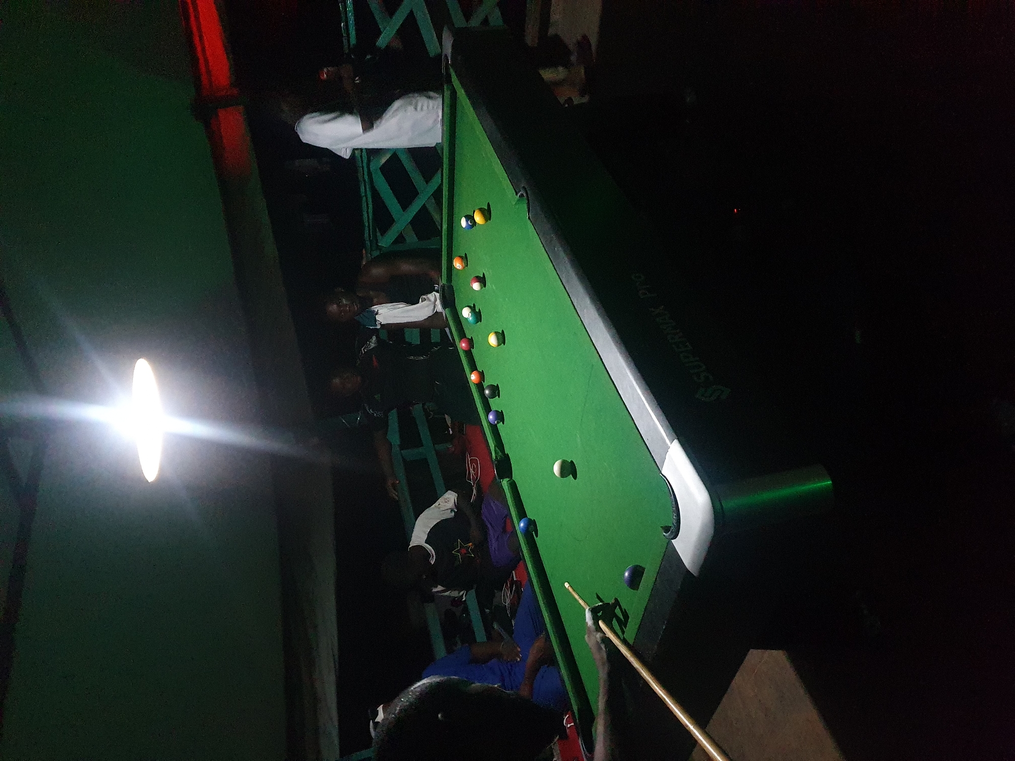 The image depicts an outdoor evening scene at a billiards table, likely in Lokoja, Nigeria, given the context. A group of individuals are gathered around a green billiards table, illuminated by a bright overhead light fixture. One person, viewed from the side, is in the process of striking a billiard ball with a cue. Other players are visible in the background, some seated and others standing. The setting appears to be a recreational area, possibly a bar or a social gathering spot. The environment suggests a warm climate, with people dressed casually. The billiards table is equipped with a full set of colored balls.