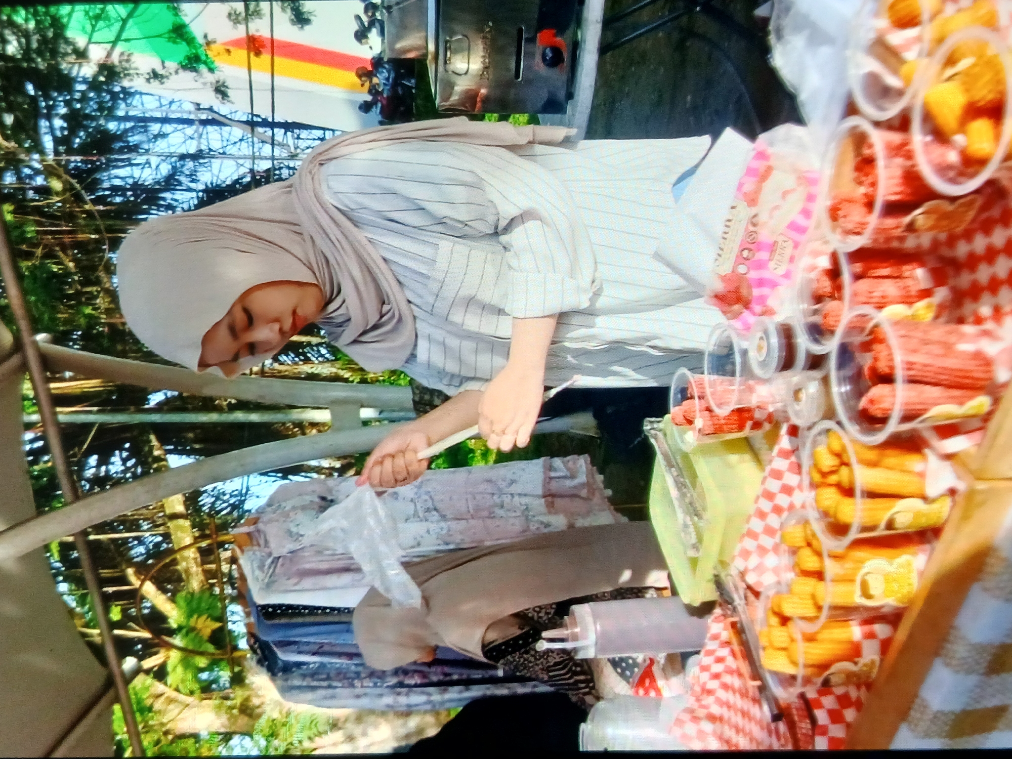 An outdoor market stall in Setia Bakti, Indonesia, features a young woman wearing a beige hijab and a striped shirt. She is in the process of packaging food items in clear plastic bags, holding a bag in her right hand and a utensil in her left. In front of her, on a checkered tablecloth, are several clear plastic cups filled with various food items, some appearing to be fried snacks like churros and others containing what look like diced fruits or candies. Behind her, a rack displays an assortment of clothing, and a food warmer is visible to the right. The background shows trees and a blue sky, suggesting a sunny day. The overall scene depicts a vendor at a bustling market.
