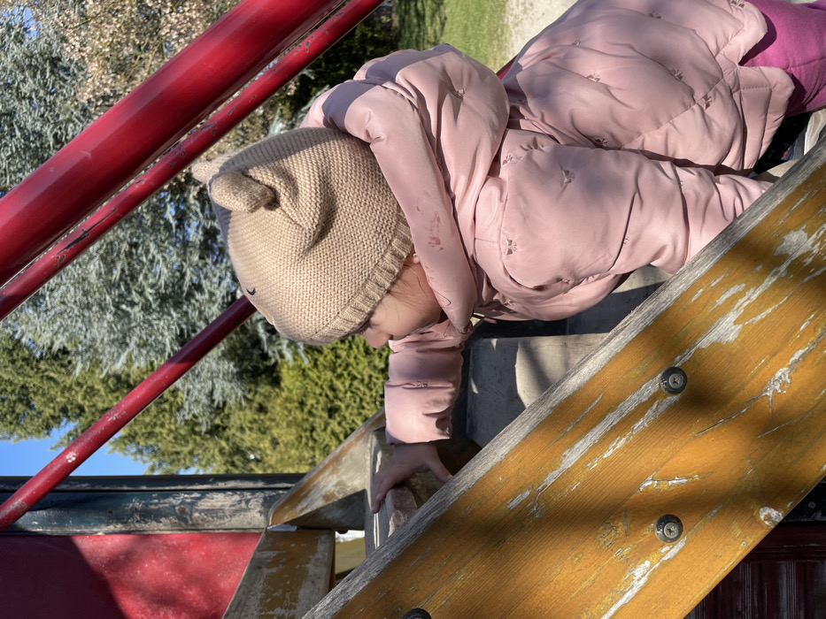 A young child is depicted outdoors on a sunny day, actively climbing a playground structure. The child, viewed in profile from the mid-torso up, wears a light brown knitted beanie with two small ear-like protrusions and a light pink puffer jacket featuring subtle embroidered patterns. A reddish stain is present on the upper left shoulder area of the jacket. The child's left hand grips a worn, light brown wooden step, which exhibits peeling paint or varnish and exposed metal screws. Reddish-orange painted metal bars, showing minor abrasions, are visible above and around the child, forming part of the playground equipment. The background consists of various green trees, including coniferous types, under a clear blue sky. A light-colored ground surface, consistent with gravel or dirt, is visible behind the child. While the setting is within Senden, Germany, the specific location or address within the city cannot be determined from the visual information provided.