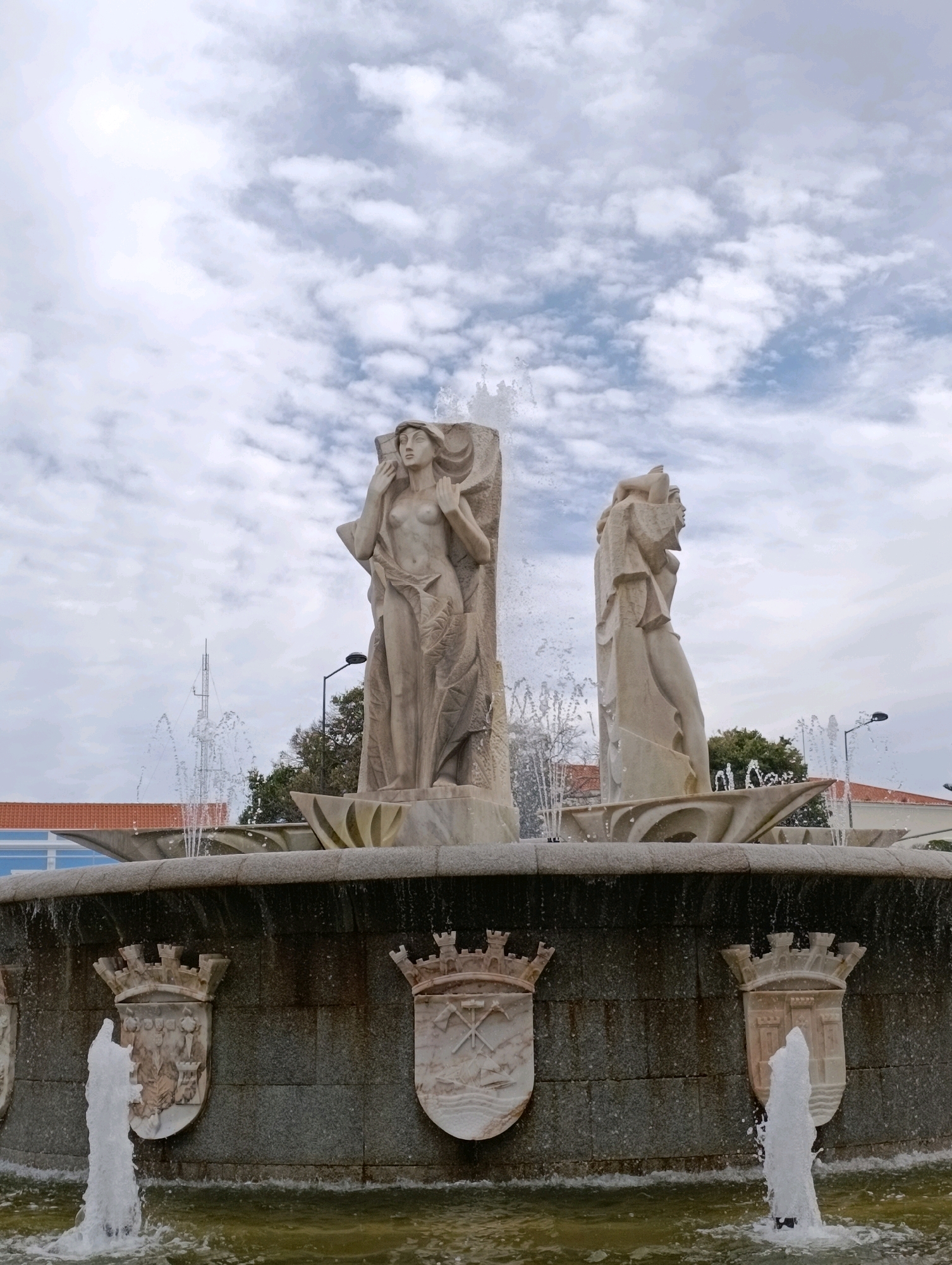 The image shows the Fountain of the Four Elements located in Praça da Liberdade, Setúbal, Portugal. The fountain features two large, beige stone sculptures of female figures, one on the left and one on the right, with water spraying from the fountain's basin. The sculptures are stylized and draped in flowing garments, with the left figure looking forward and the right figure in profile. The fountain's basin is made of dark stone and is adorned with several heraldic crests. Water jets spray from various points in the fountain, creating a dynamic effect. In the background, a cloudy sky with patches of blue is visible, along with trees, a lamppost, and parts of buildings. The overall scene depicts a public monument in an urban setting.