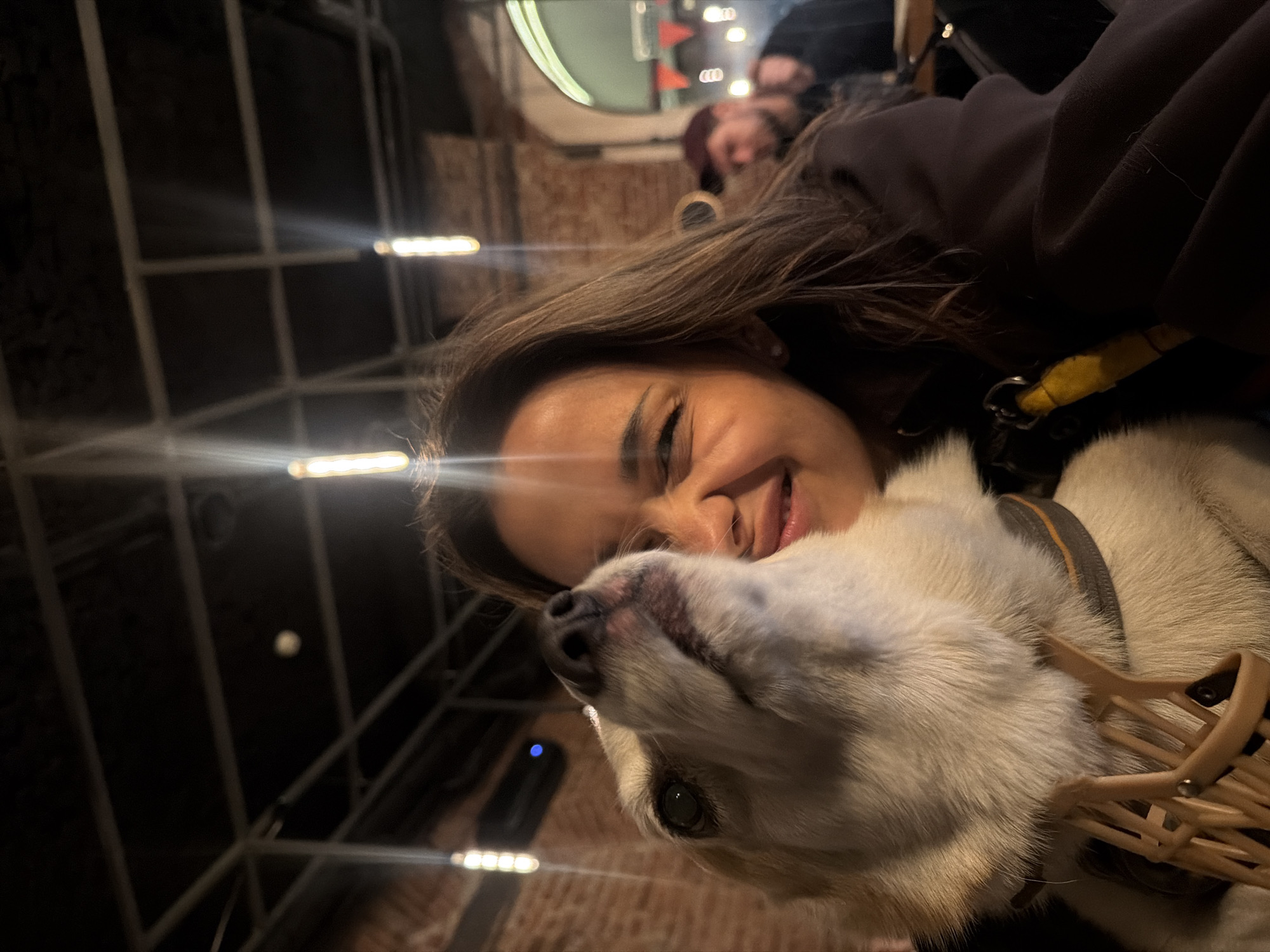 The image shows a close-up of a woman and a dog in a dimly lit indoor setting, possibly a bar or restaurant in Saint Petersburg, Russia. The woman is smiling, with her eyes closed, and her head is tilted back slightly. The dog, a light-colored breed, is looking up and to the left, with its mouth open. The dog is wearing a muzzle and a collar. In the background, out of focus, a man wearing a baseball cap can be seen. The environment appears to be industrial or rustic, with exposed brick walls and hanging light fixtures.