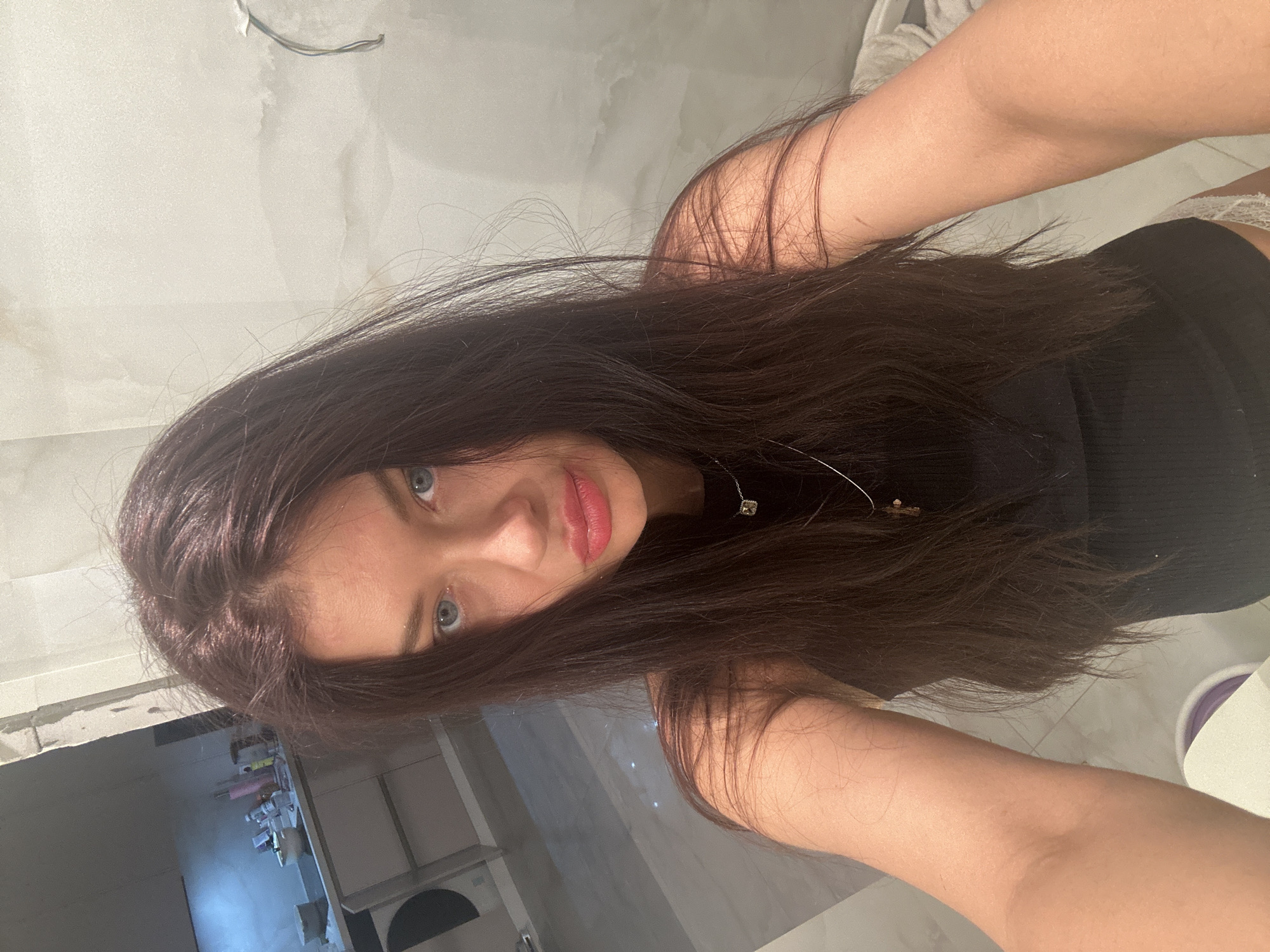 A young woman with long, dark brown hair and striking light blue eyes looks directly into the camera, capturing a selfie in an indoor setting. Her head is tilted slightly, and her hair falls forward, partially obscuring her face. Her lips are full and tinted pinkish-red, slightly parted. She has fair skin and a neutral, engaging expression.
She is wearing a dark, possibly black, ribbed top, and a delicate silver necklace with a small, light-colored pendant is visible on her chest. Her arms are raised and visible on either side of her, indicative of holding a phone or camera to take the picture.
The background suggests an unfinished or undergoing renovation space. Light-colored walls, possibly off-white or light grey, appear textured or uneven. Exposed wires hang from the ceiling in the upper left, reinforcing the construction theme. The floor, partially visible in the lower right, is tiled with light grey or white squares. On the left side of the frame, there's a cluttered area with various items and a bright light source. On the right, parts of white and dark objects, possibly furniture or appliances, are visible. A small white, possibly lace, detail peeks out near her right arm. The overall scene conveys an unkempt or transitional environment, possibly within a home or apartment in Moscow, Russia. There is no visible text.