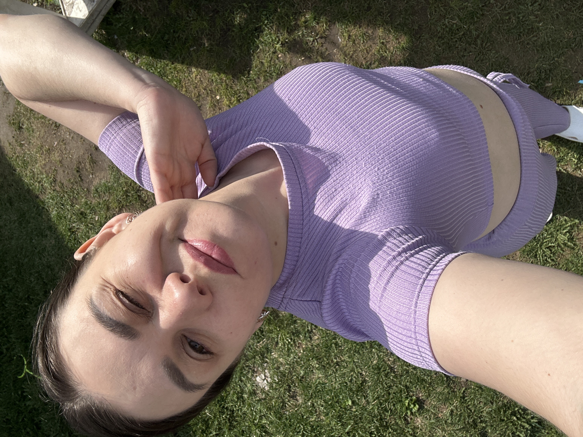 A woman is lying on her back in a grassy area, likely outdoors, based on the presence of green grass. She is wearing a lavender, ribbed, two-piece outfit that reveals a strip of her abdomen. The photograph appears to be a selfie, as one arm is extended, presumably holding the camera. She is partially squinting with one eye closed. The location is purportedly in Chaikovsky, Russia, though there are no visible clues in the image to confirm this. The woman is wearing earrings. There are pieces of wood visible in the background.