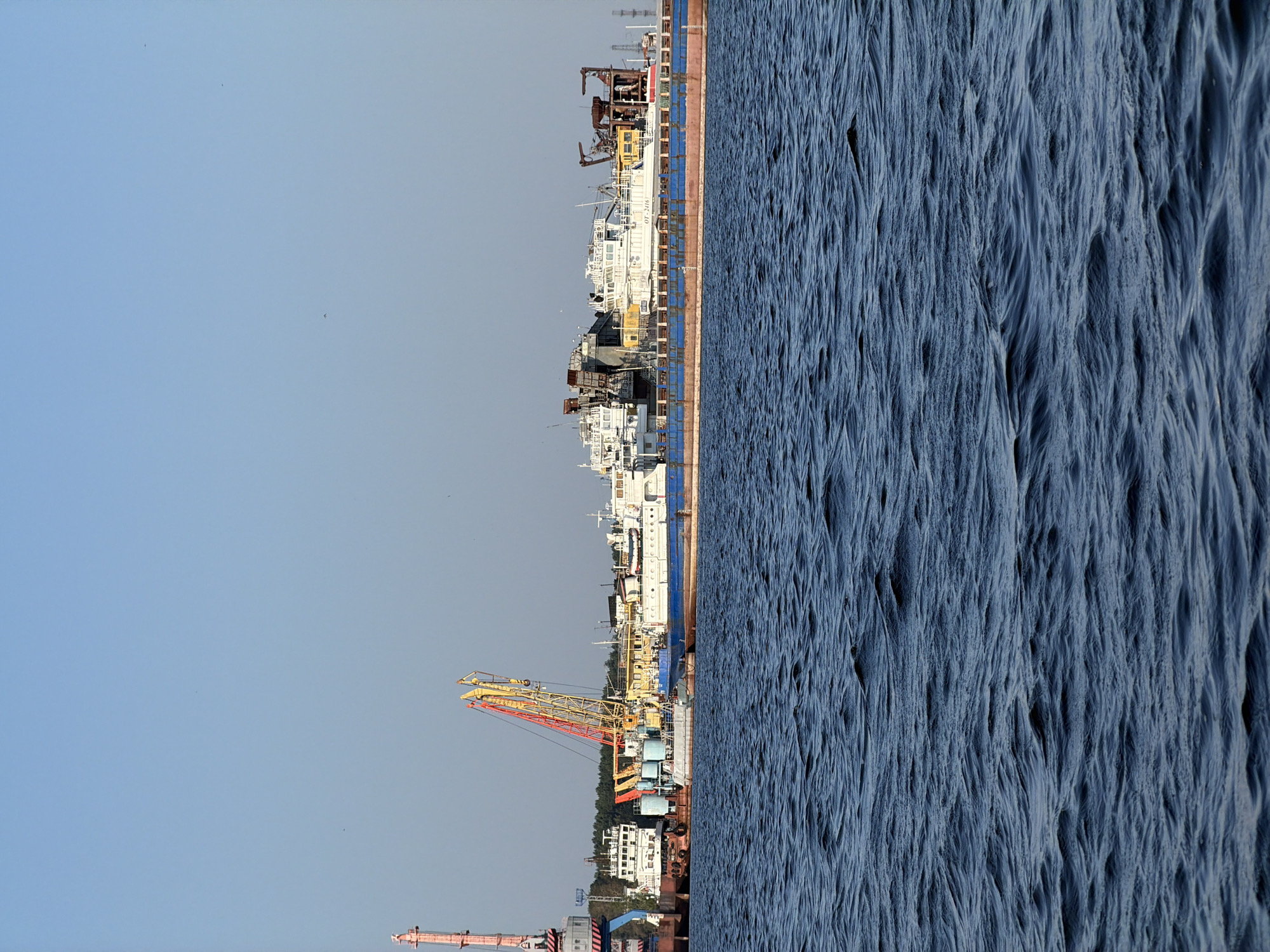 A wide shot captures a view from the Kama River in Chaikovsky, Russia, looking towards a shipyard under a clear, bright blue sky. In the foreground, the dark blue water of the river is visible, with small, choppy waves. The middle ground is dominated by a series of docked ships and industrial structures. A large, long barge with a blue hull and a rust-colored upper section is prominent. On top of this barge are several white vessels, some appearing to be research or work boats, with complex superstructures and cranes. To the left, a large pink and grey industrial crane stands tall. Yellow and red cranes are also visible, extending over the ships. In the background, a dense line of green trees forms a backdrop against the horizon, with some distant industrial towers. The scene suggests ongoing activity at a shipyard or port.