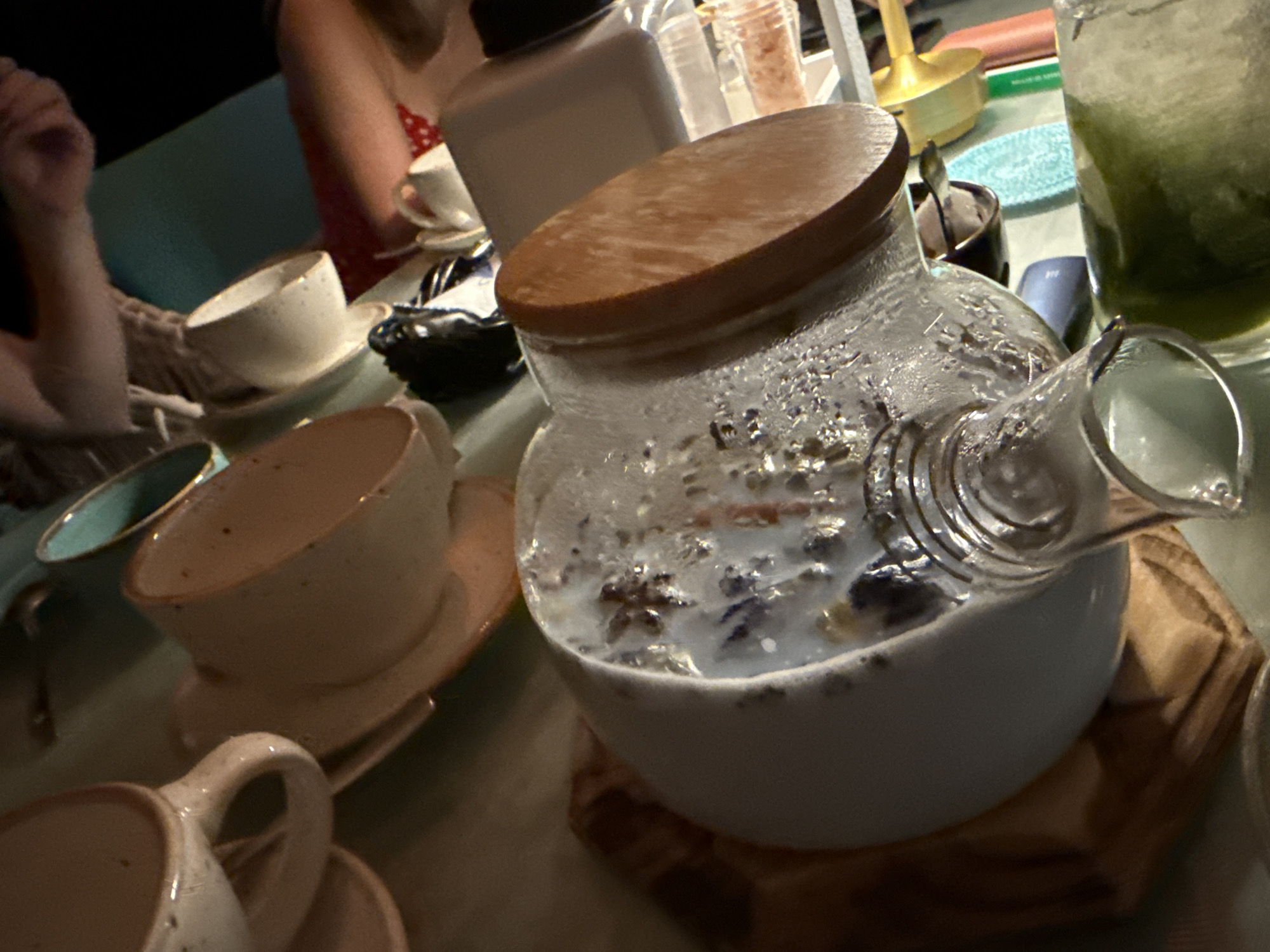 A close-up, indoor, eye-level shot captures a table laden with tea-related items. In the foreground, a light-colored ceramic teapot with a wooden lid holds a milky beverage with what appear to be flower petals or herbs. To its left, a light-colored ceramic cup and saucer are visible. Further left, a teal-colored shallow dish with a gold rim sits on the table. Behind these, a larger, light-colored ceramic cup and saucer is positioned. In the background, out of focus, a person wearing a red and white polka-dot dress is visible, along with other objects including a clear glass containing a green beverage, a salt shaker, and a white container. The lighting is dim, suggesting an evening or dimly lit interior. The location is identified as Feodosia, Ukraine.