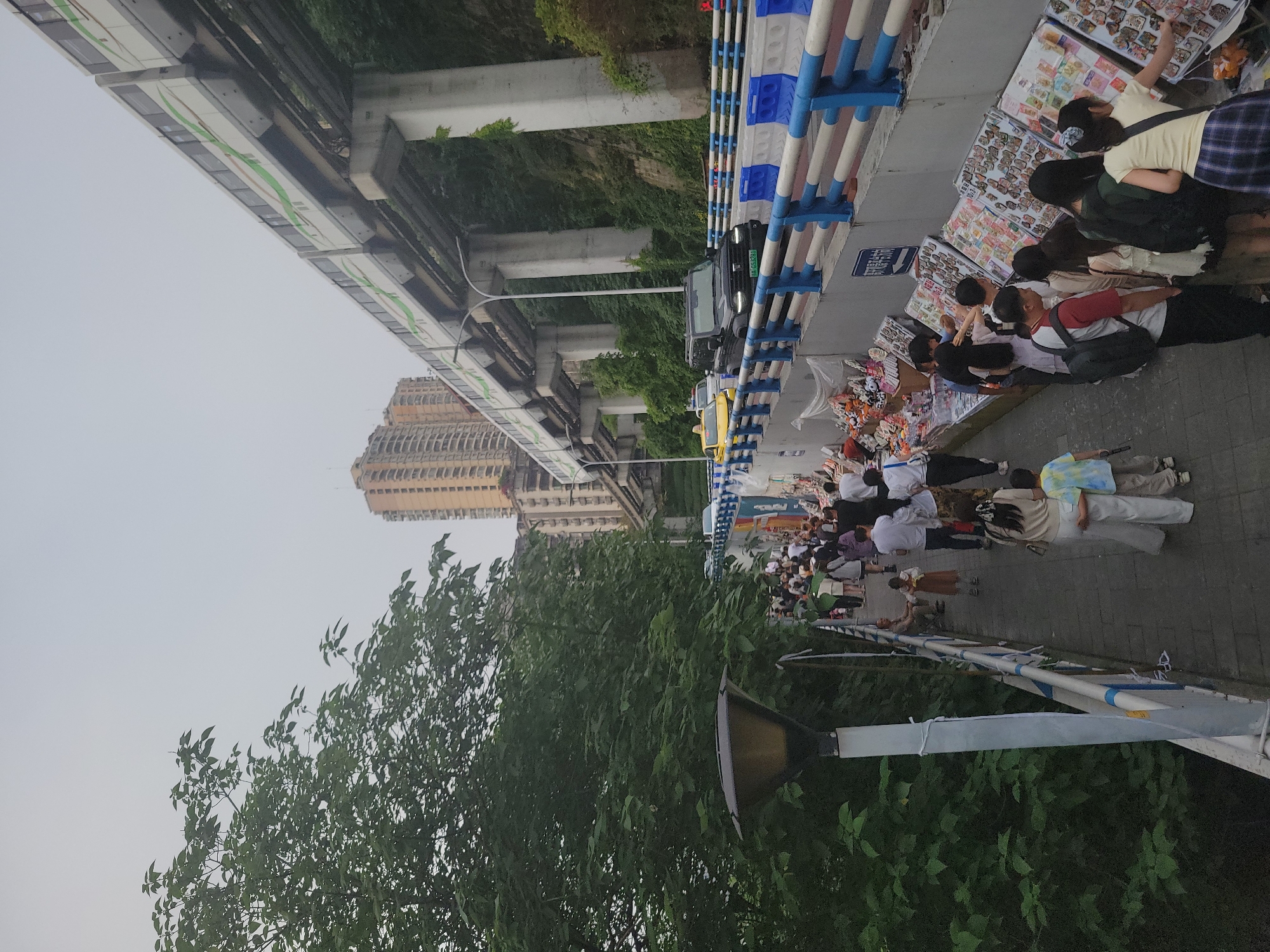 The image depicts a bustling scene on a sidewalk in Yuzhong District, Chongqing, China. People, primarily young, are gathered along a row of vendor stalls displaying souvenirs and trinkets. An elevated railway line runs parallel to the sidewalk, supported by concrete pillars. Cars can be seen on the road adjacent to the sidewalk. The architecture visible includes a tall apartment building in the background. Lush green trees and foliage are prominent in the foreground. The weather appears overcast. The sidewalk is lined with blue and white safety barriers.