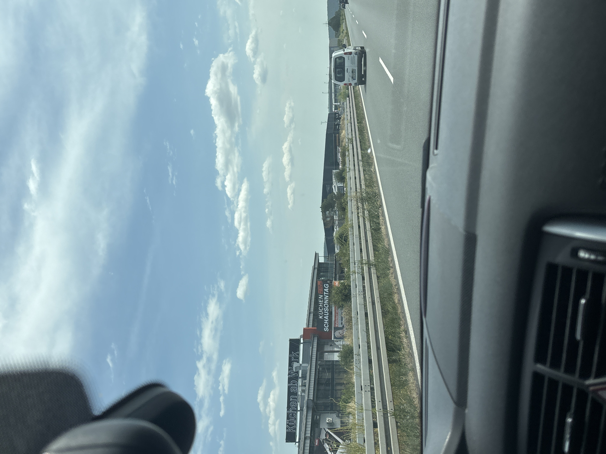 The image captures a view from inside a vehicle traveling on a highway. The foreground shows the dashboard and the lower portion of the windshield. Beyond the highway guardrail, a commercial building is visible with signs reading "Küchen ab Werk" and "KÜCHEN SCHAUSONNTAG". Further in the distance, several other industrial buildings and a white van are seen on the highway. The sky is blue with scattered clouds. This location is the city of Großwallstadt, Germany.