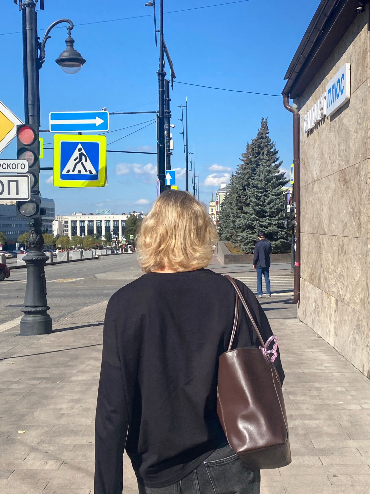 A person with blonde hair is walking away from the camera on a sidewalk in Perm, Russia. The person is wearing a black long-sleeved shirt and dark jeans, and carrying a brown leather bag with a pink charm. In the background, there is a street with a pedestrian crossing and traffic lights. A man in a dark jacket and jeans is standing at the crossing. The sidewalk is made of paving stones, and there are trees and buildings visible in the distance. The sky is blue with some clouds. A sign on a building reads "ЦУМ ПЛЮС".