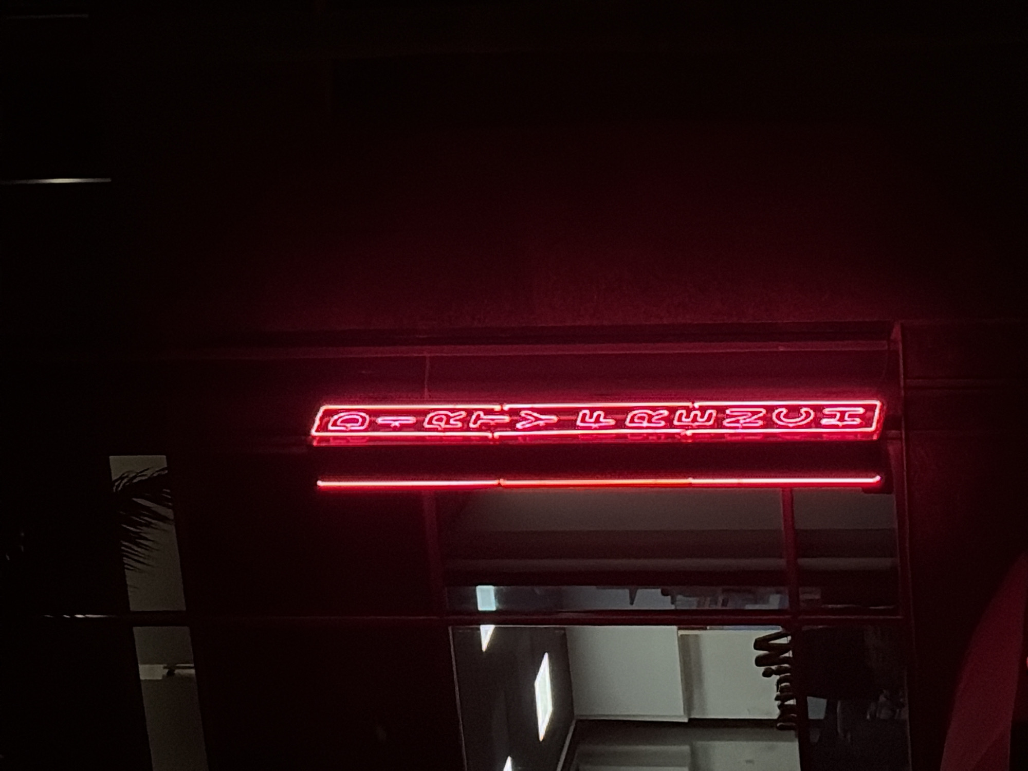 The image shows a neon sign reading "DIRTY FRENCH" in red, possibly the entrance to a restaurant. The location appears to be in an urban setting, possibly in Miami, Florida, judging by the architectural style of the building and the presence of a palm tree silhouette visible through a window. The sign is positioned above an entrance, with reflections of the interior visible. The lighting is dim, creating a dark and moody atmosphere.