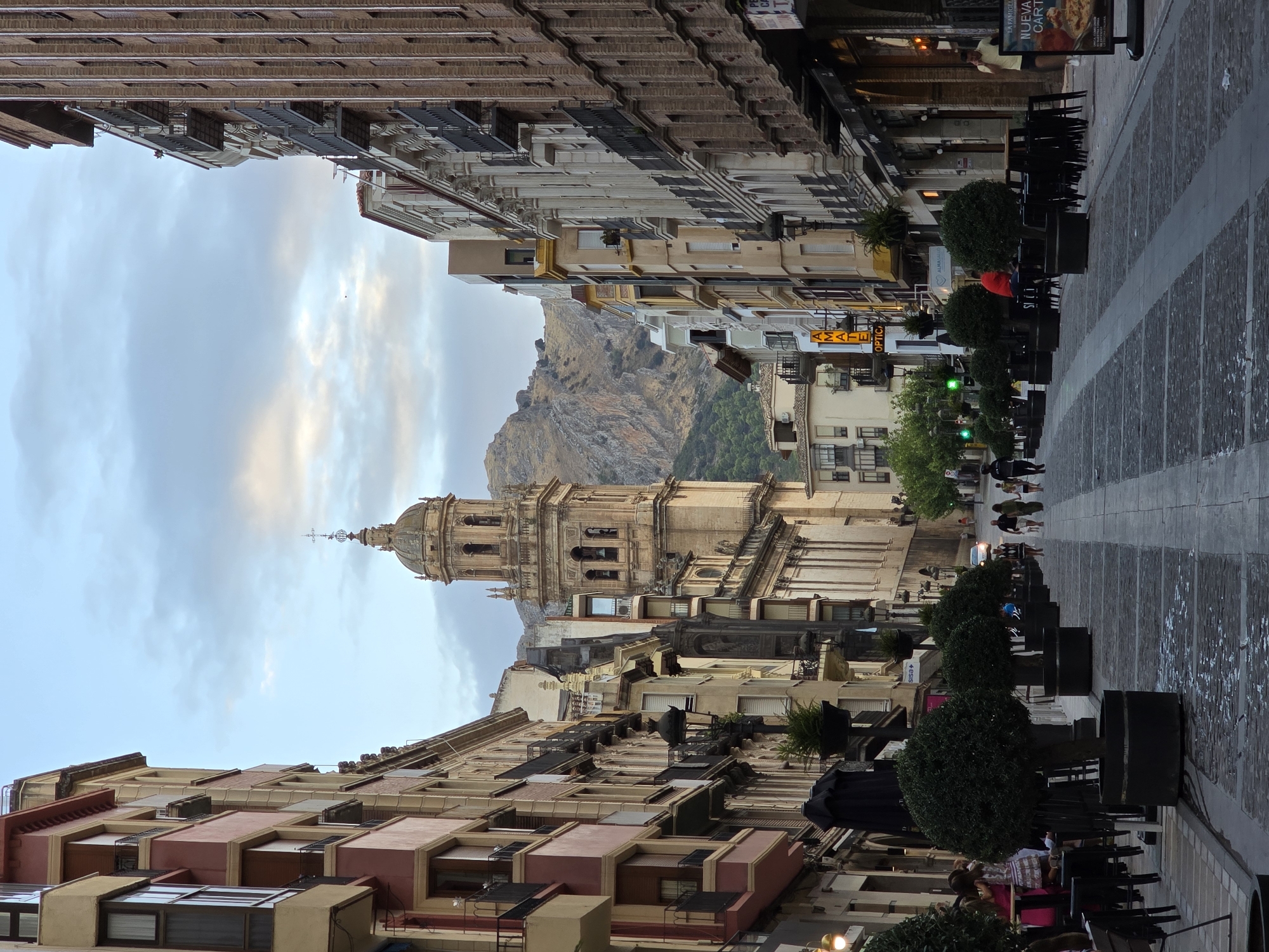 The image depicts a view looking down a wide, stone-paved pedestrian street lined with multi-story buildings on both sides. In the distance, a prominent bell tower of a cathedral or large church is visible, with a steep, rocky mountain behind it. The sky is overcast with scattered clouds.
On the left side of the street, the buildings are a mix of architectural styles, with some featuring balconies and ornate facades. There are trees in large planters along the sidewalk, and outdoor seating areas for cafes or restaurants. Several people are visible walking down the street, some with shopping bags, and others sitting at tables.
On the right side of the street, similar buildings with shops at ground level face the street. The architecture here also includes balconies and brickwork. More outdoor seating is present, and a few individuals are seen walking.
The overall impression is of a bustling European city street, likely in a historic district, with a blend of commercial activity and pedestrian traffic. The presence of the cathedral and the mountain in the background suggests a city with a significant historical and natural landscape. Based on the architectural style, the bell tower's design, and the mountain backdrop, the location is likely Jaén, Spain. Specifically, the view is down Calle Maestra towards the Iglesia de la Magdalena and the backdrop of Sierra Morena mountains.