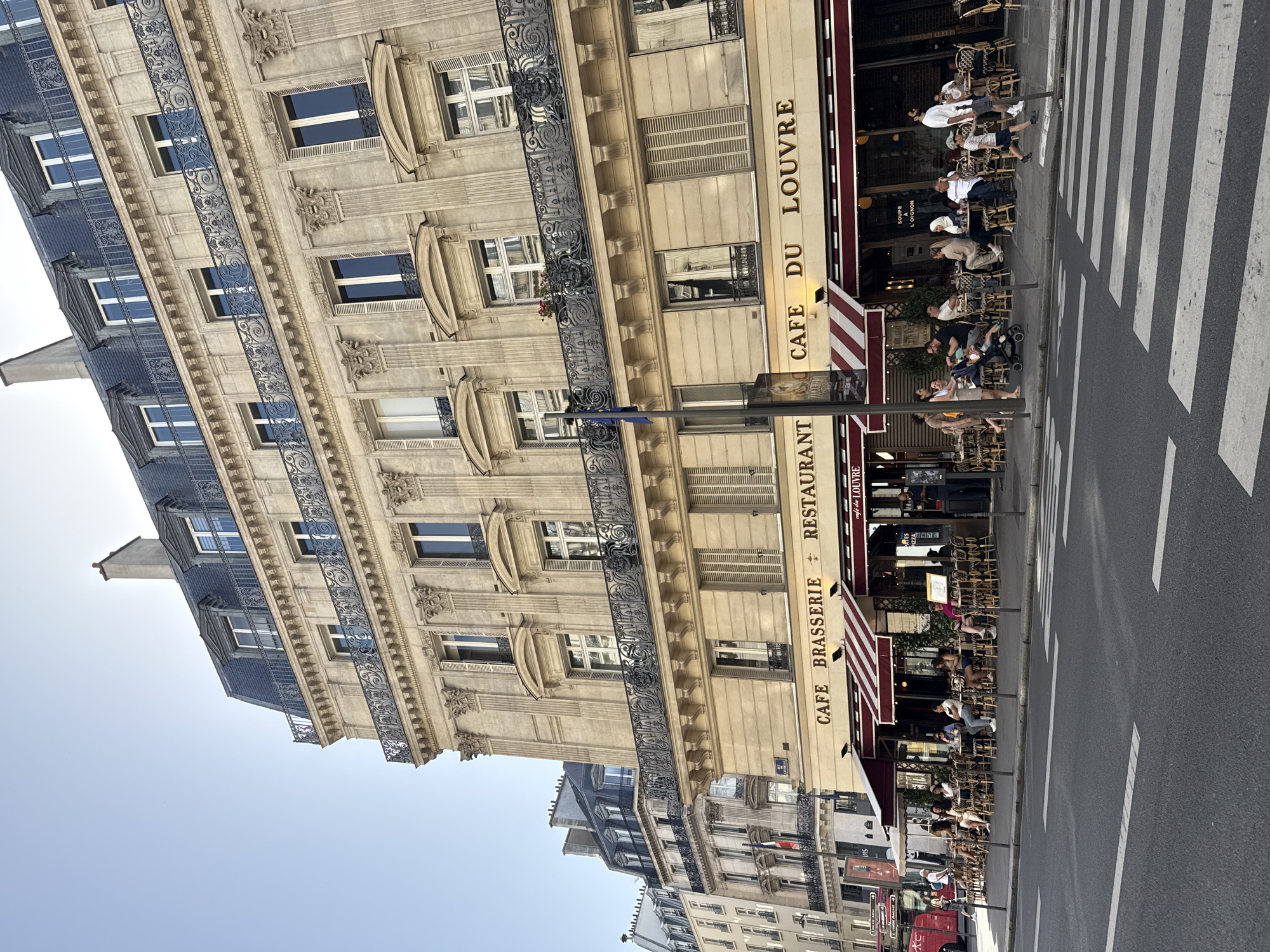 The image depicts a street scene in Paris, France, showing the facade of a building with the "Cafe du Louvre" brasserie and restaurant. The architecture is typical Parisian, with ornate windows, wrought iron balconies, and a multi-story structure. Patrons are seated at tables outside the cafe, engaging in typical cafe activities like eating, drinking, and socializing. A pedestrian crosswalk is visible in the foreground, suggesting a busy urban environment. The sky is clear. Other buildings are seen down the street, indicating a continuation of the urban setting.