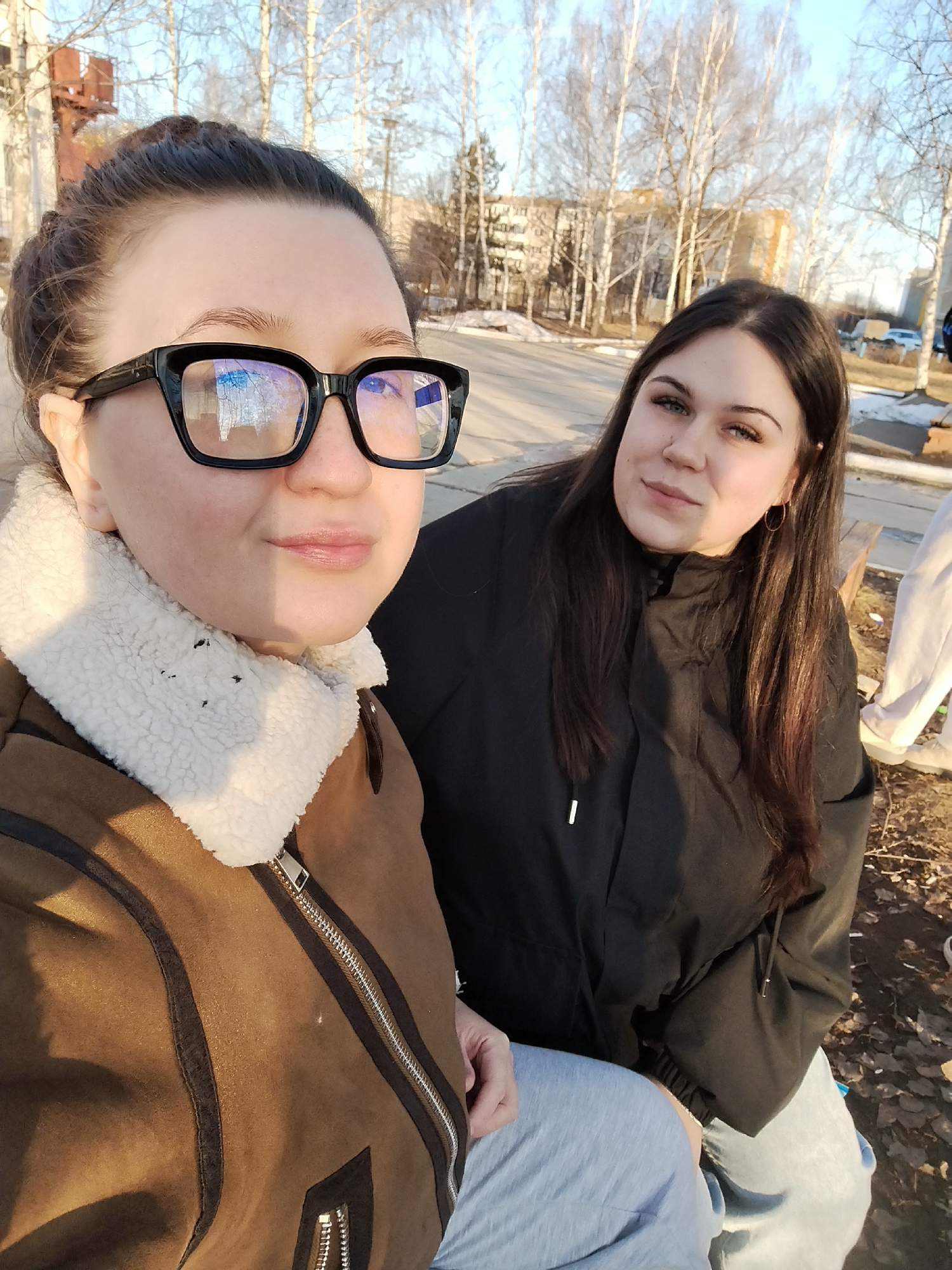 Two women are posing for a selfie outdoors. The woman on the left is wearing a brown jacket with a white fur collar and black-framed glasses. The woman on the right is wearing a black jacket. They both appear to be sitting down.
Behind them, the scene is a paved area with bare trees, suggesting either late fall or early spring. There is a building in the background and some patches of snow on the ground. The location is the city of Chaikovsky, Russia.