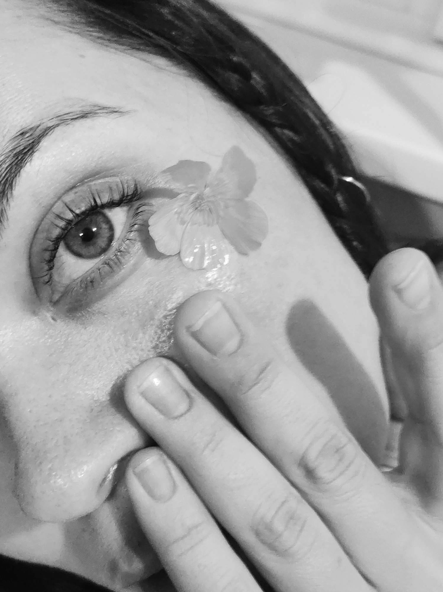 This black and white, close-up portrait depicts a portion of a human face, specifically the right eye, eyebrow, upper cheek, and nose, partially obscured by a hand. A small, delicate flower with five petals rests on the upper cheekbone directly beneath the outer corner of the eye. The eye is open, revealing a detailed iris and pupil, framed by distinct eyelashes, which appear coated. A well-defined eyebrow is visible above the eye. A hand, positioned with fingers partially curled, covers the lower part of the nose and adjacent cheek. The fingernails are visible on the fingers, which show natural luster. Skin texture, including pores, is discernible across the face. A dark, braided segment of hair is visible on the upper right side of the frame. The lighting creates soft shadows and highlights, emphasizing contours.
Location: Undiscernible, specified as Unknown, Unknown.