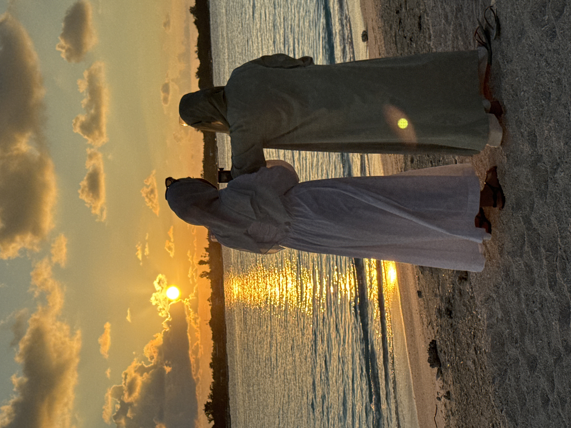 Two individuals in draped robes stand on a beach at sunset. The individual closer to the camera is wearing a light-colored robe, while the other individual is wearing a darker-colored robe. The sun is setting over the water, reflecting on the surface. The location is likely near the city of Unknown, Indonesia, as specified. The individuals are standing on the sand, near the water's edge.